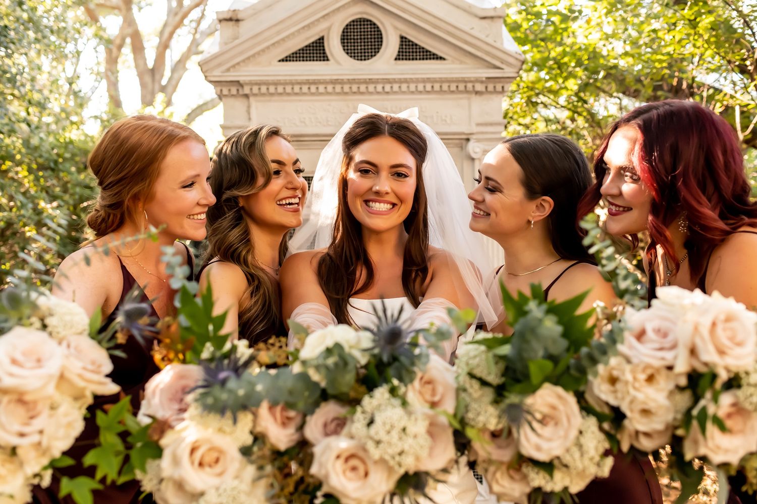 A bridal party shares joyful laughter together while holding beautiful peach and cream rose bouquets outdoors.