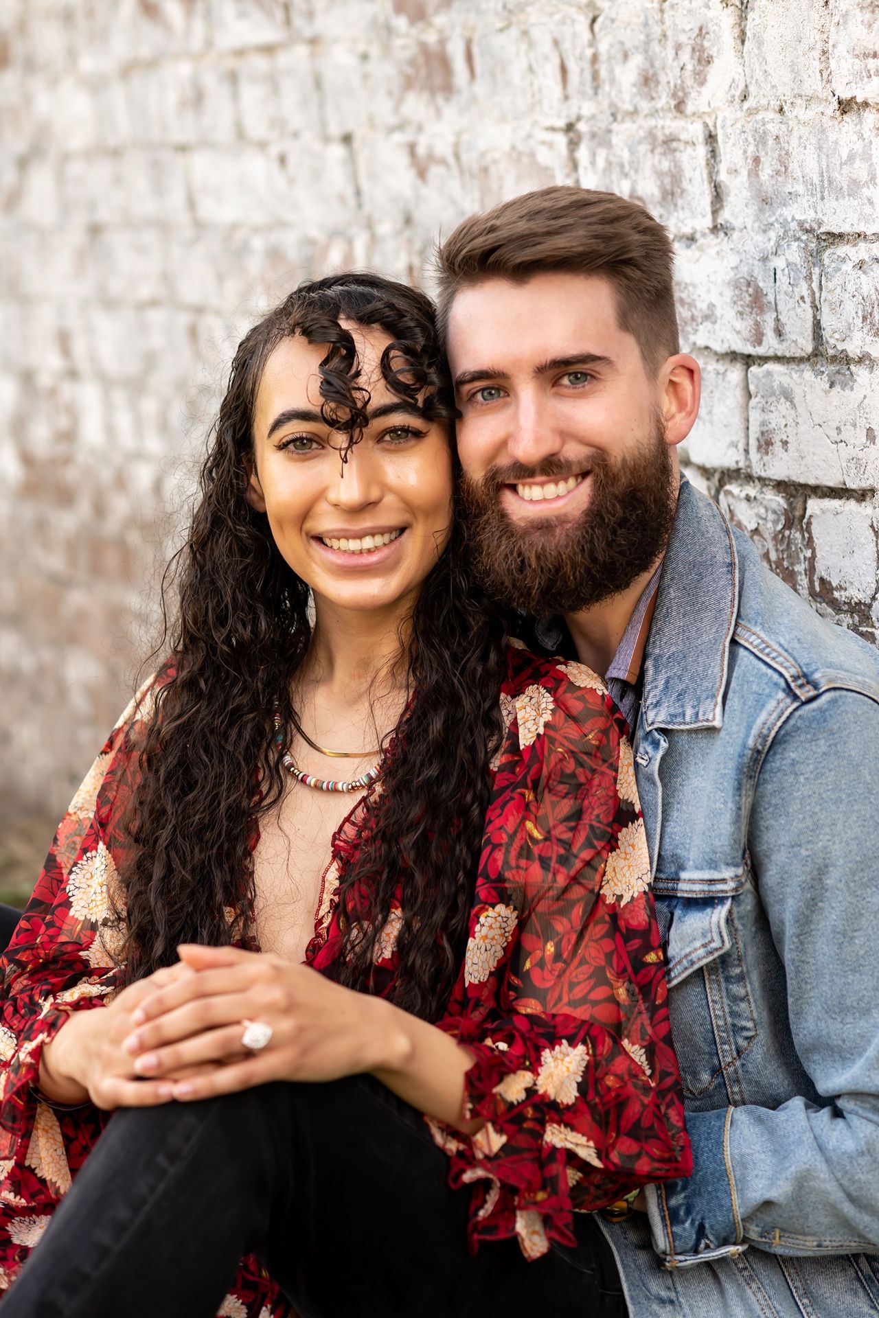 A couple in casual attire poses against a brick wall, wearing a floral dress and denim jacket.