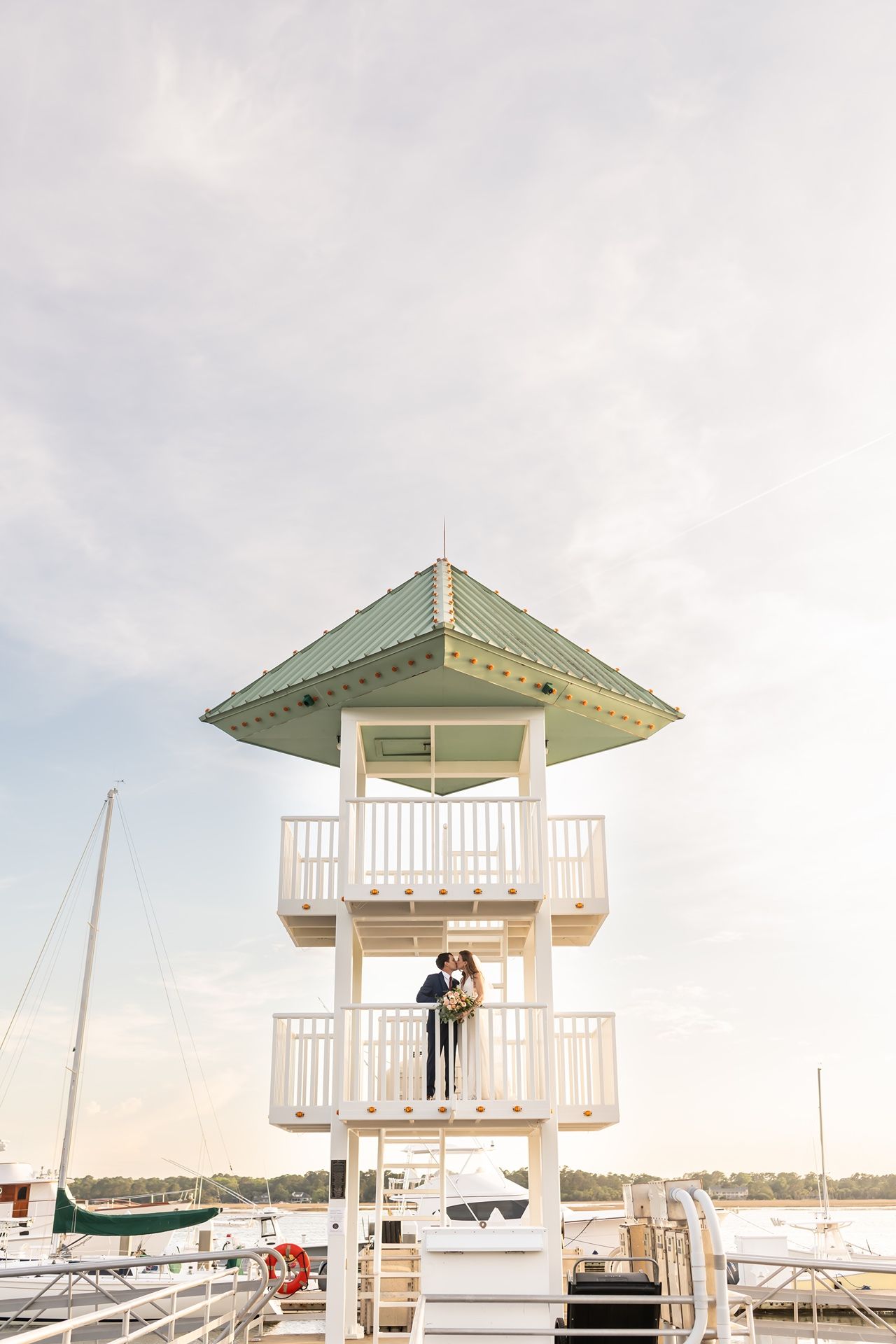 A charming white lighthouse-style lookout tower stands at a marina with boats docked in the tranquil water at sunset.