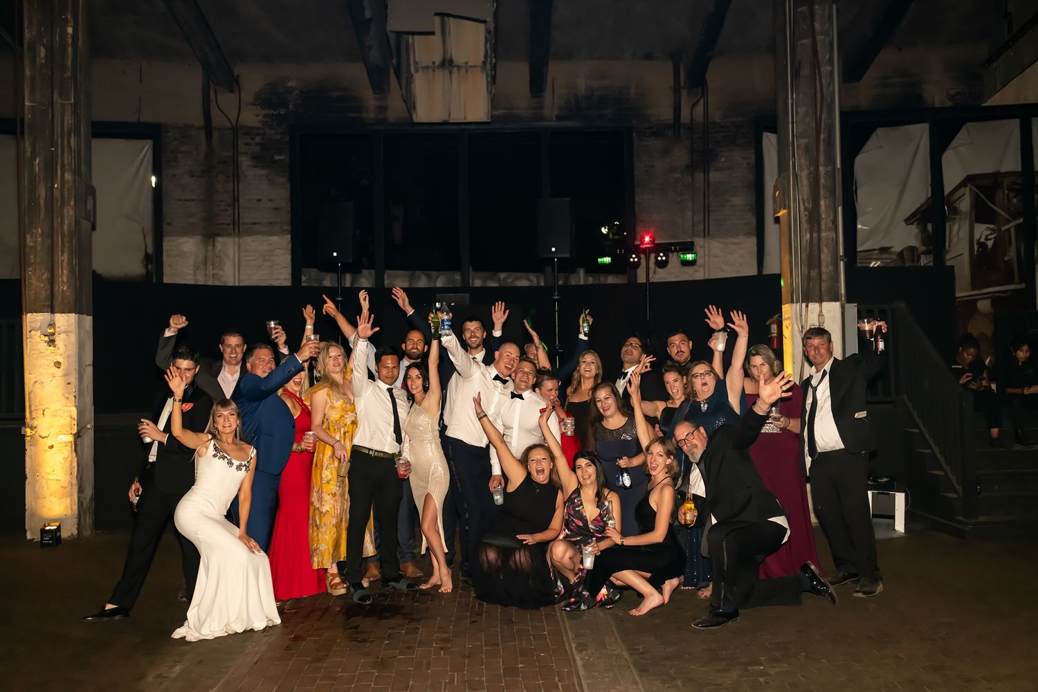 Wedding guests dancing and celebrating together at an indoor reception venue with dark atmospheric lighting.
