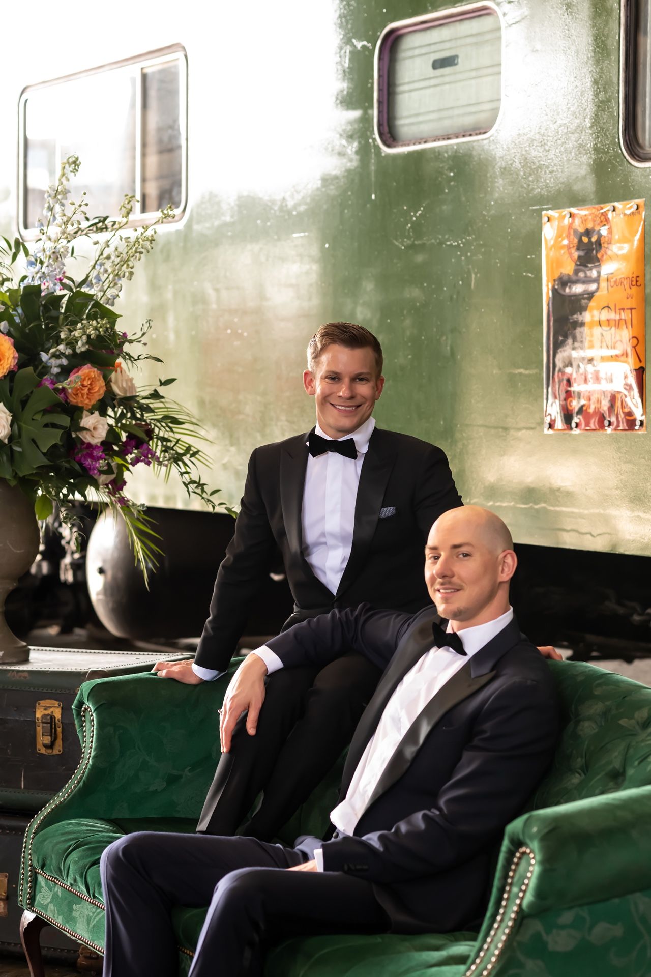 Wedding couple lounging on green velvet seats in a vintage train car decorated with floral arrangements.