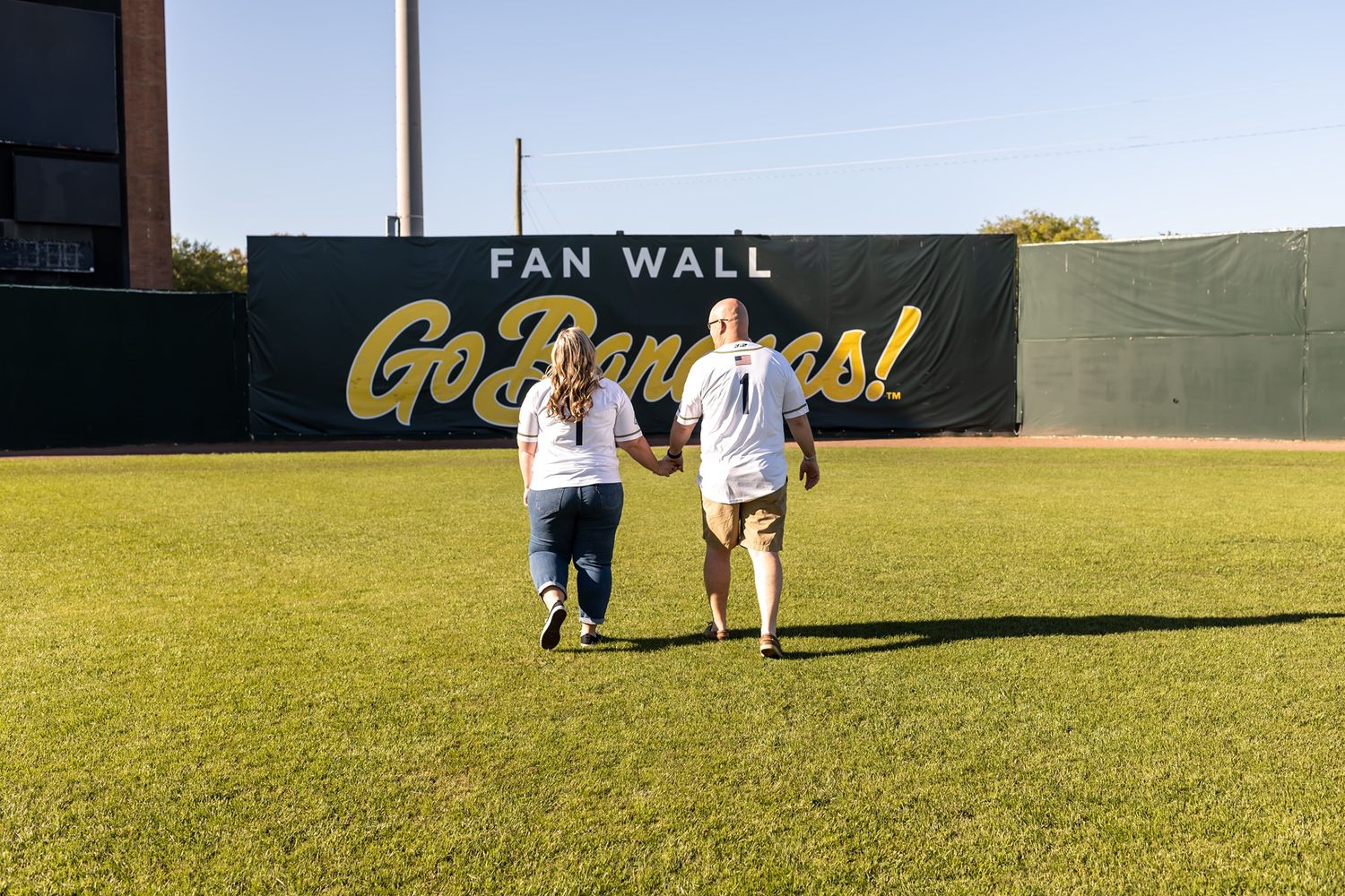 Two people walking across the outfield grass at a baseball stadium, with the iconic Fan Wall in the back.