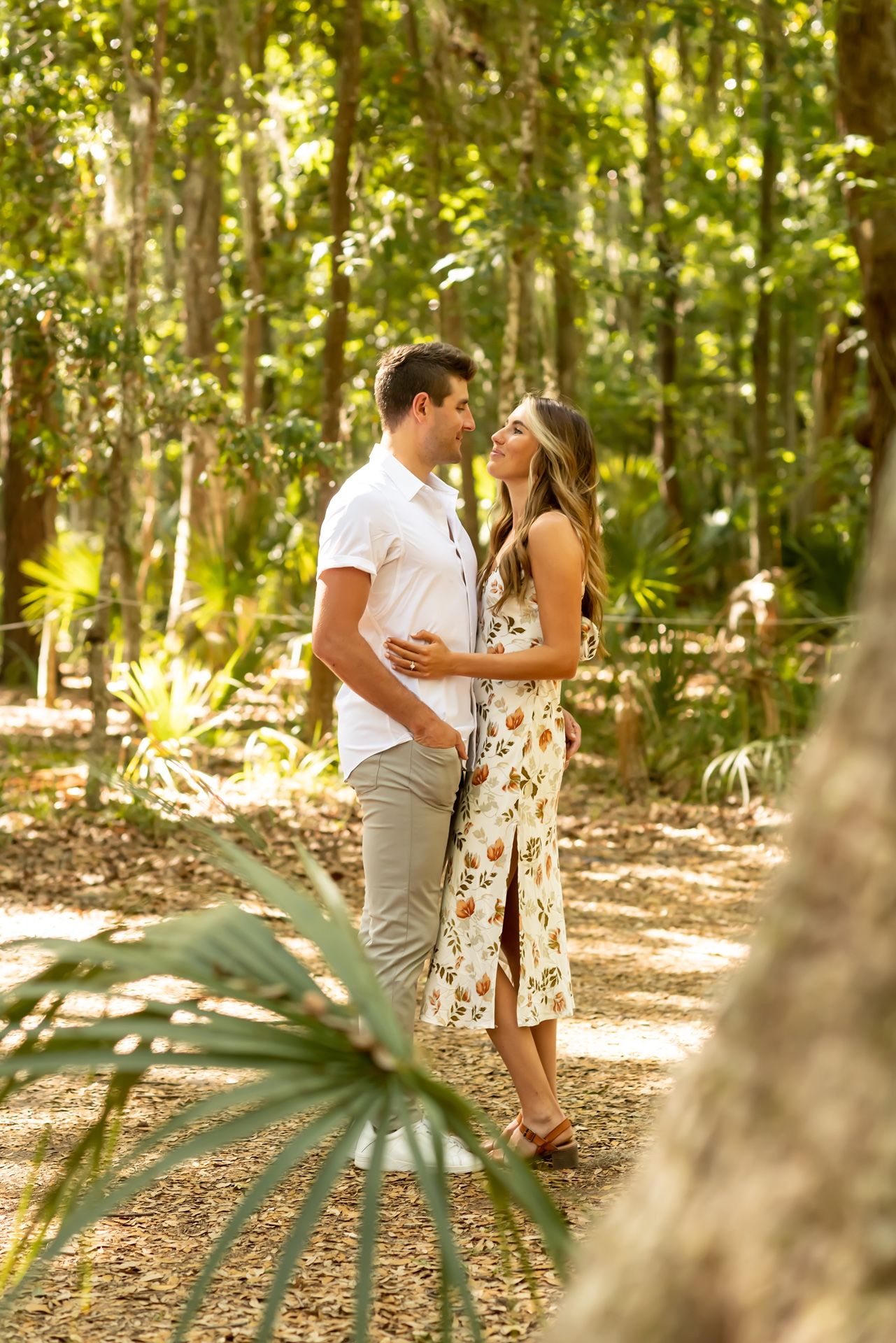Couple embraces lovingly in a sunlit forest clearing with soft golden light filtering through trees.
