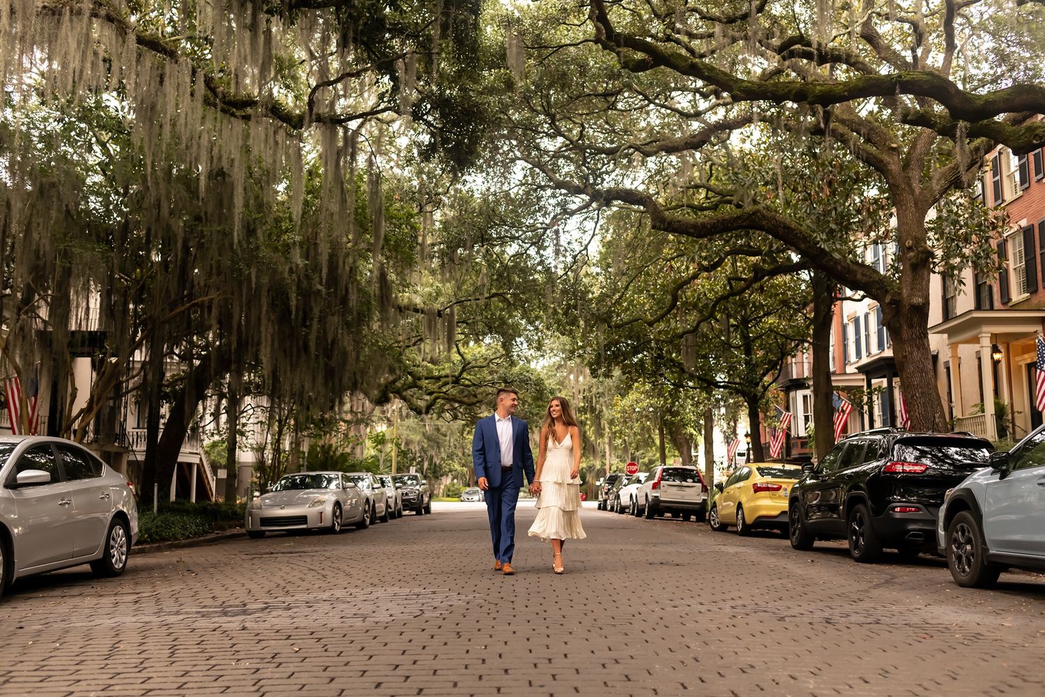 Tree-lined cobblestone street in historic Savannah with Spanish moss hanging from oak branches and parked cars along sidewalks.