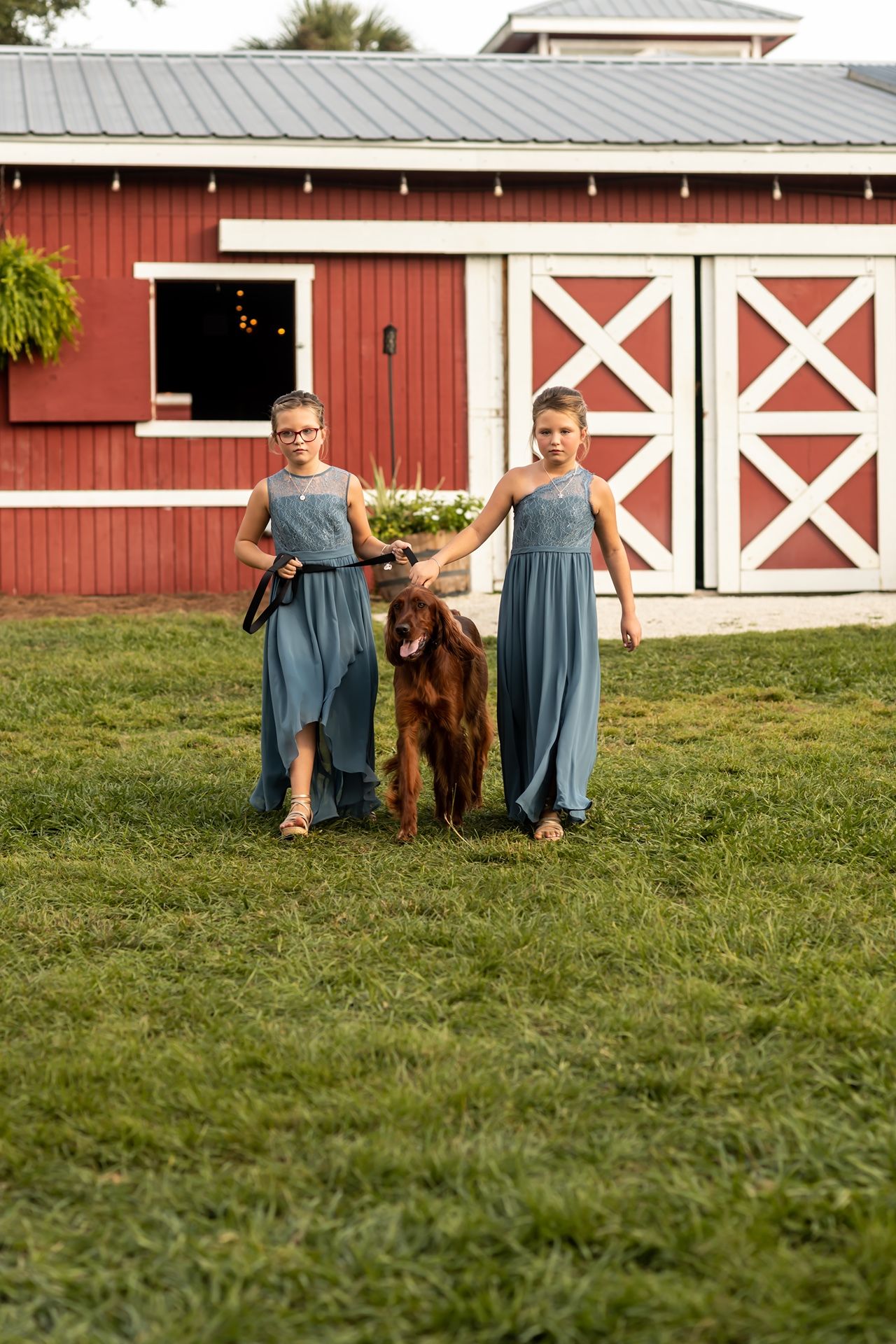 Two people in matching blue dresses stand with a brown horse in front of a red barn with white X doors.