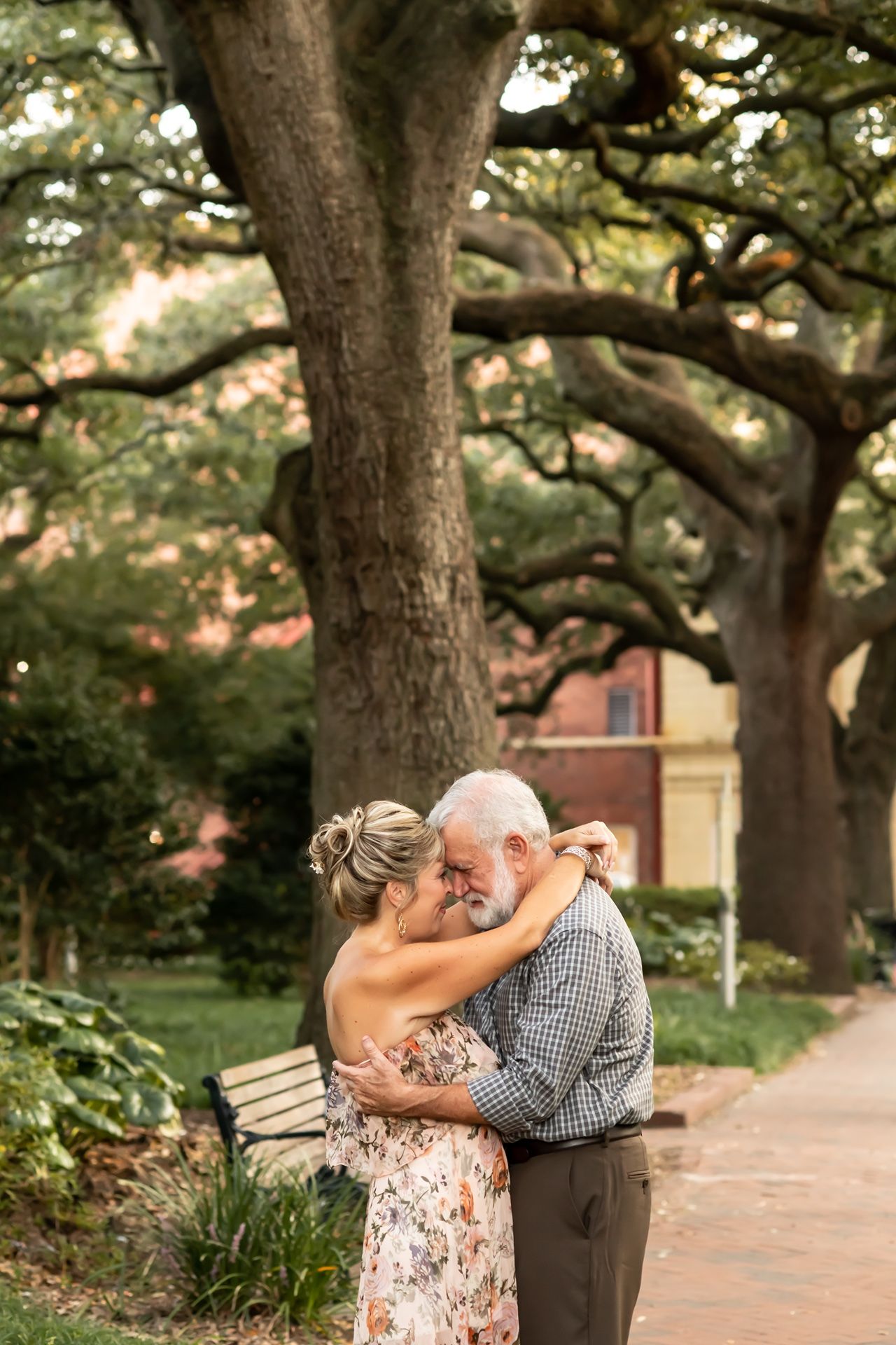 A couple shares an intimate moment together under large oak trees on a brick path.