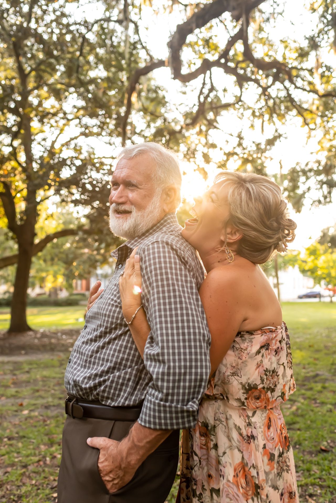 A floral-dressed woman and plaid-shirted man in a loving outdoor embrace with golden sunlight filtering through trees.