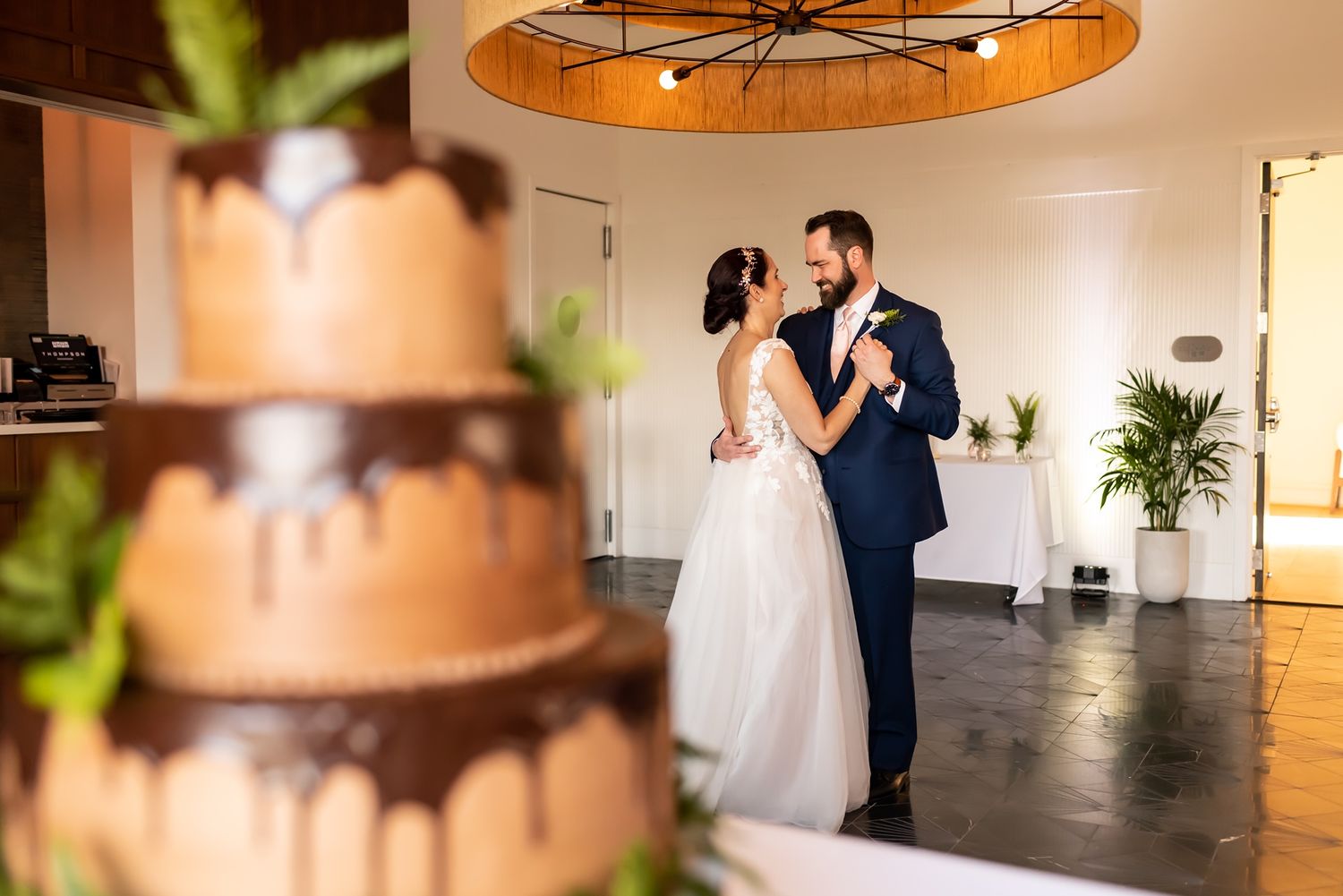 A romantic wedding scene with a couple dancing next to an elegantly tiered chocolate cake under a modern pendant light.