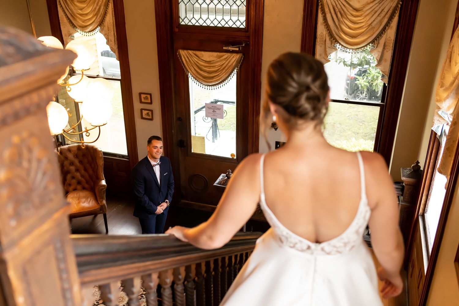 A bride in a white wedding dress stands at the top of an elegant staircase in a historic venue with ornate decor.