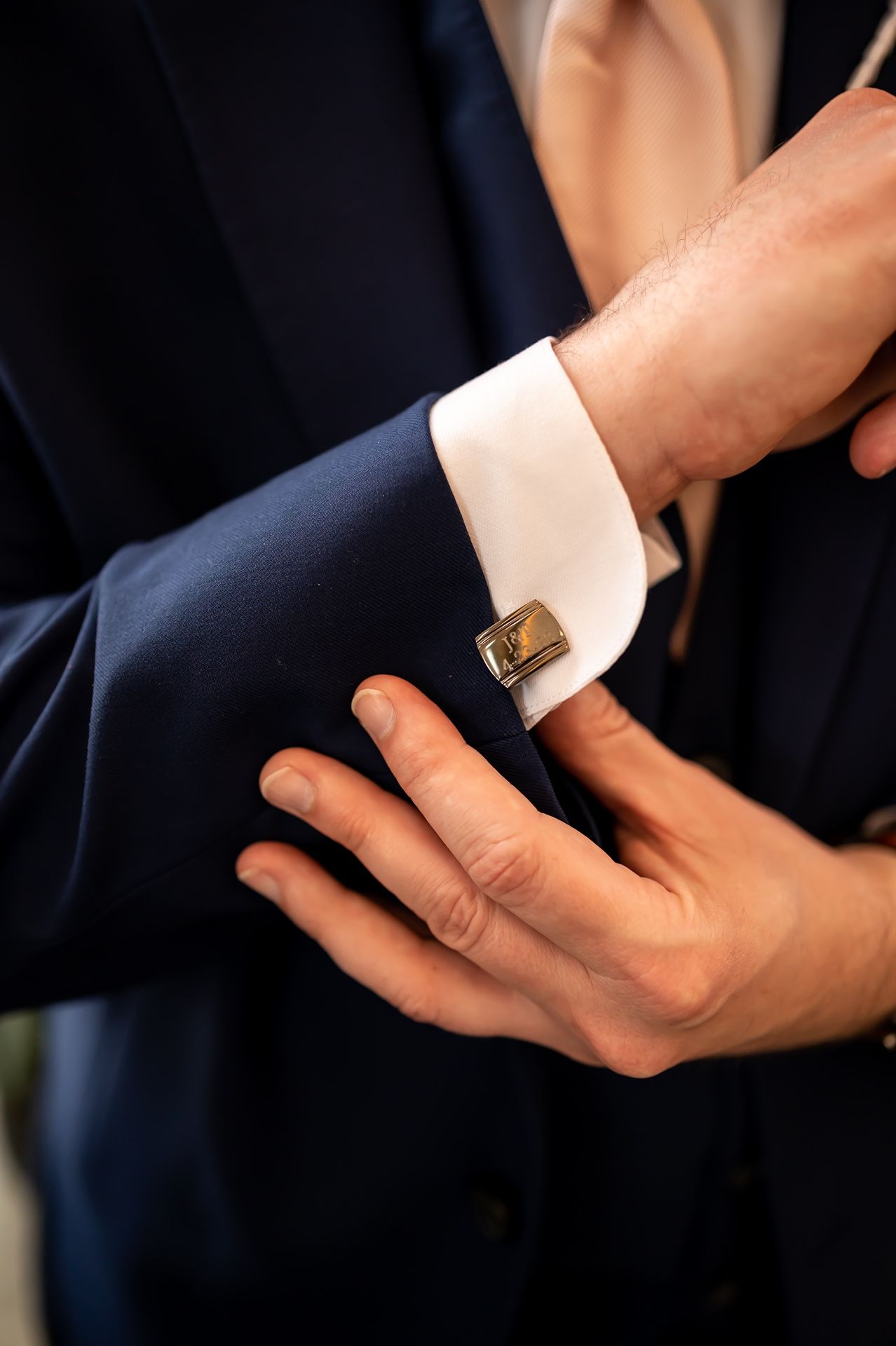 Close-up shot of hands adjusting a gold cufflink on a white dress shirt sleeve with navy blue suit jacket.