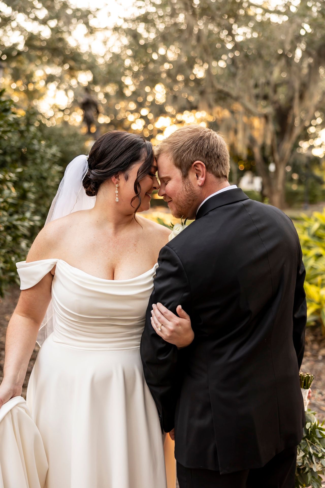 Romantic newlywed couple shares an intimate moment in a garden at sunset with glowing bokeh lights behind them.