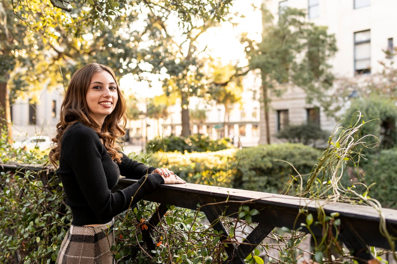 A woman sits working outdoors at a desk against a backdrop of lush greenery and sunlit trees in an urban setting.