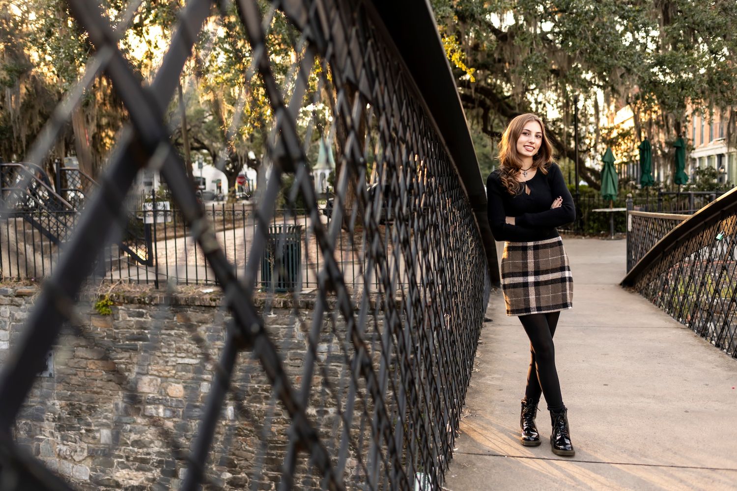 A figure in a plaid skirt poses on a stone bridge lined with black metal railings during golden hour lighting.