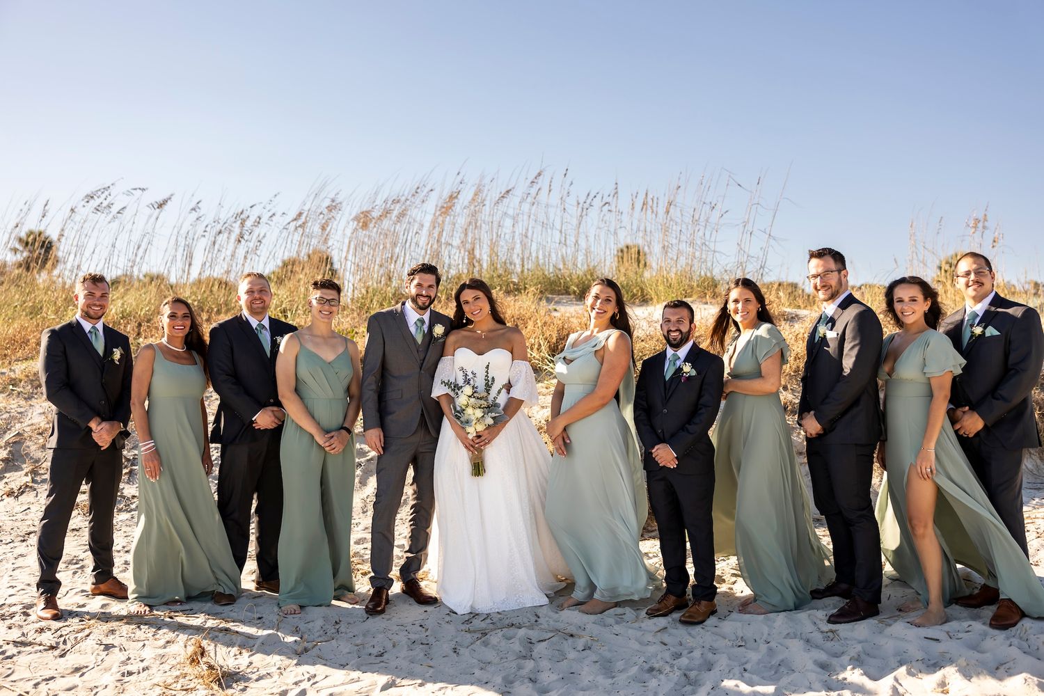 Wedding party in sage green bridesmaid dresses and dark suits pose together on sunny beach with sea grass.