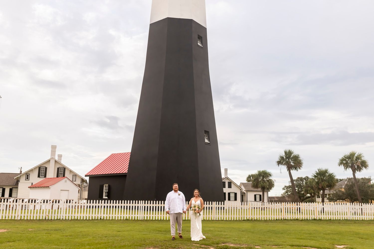 A historic black lighthouse with red-roofed buildings and white picket fence stands against a cloudy sky at Tybee Island.