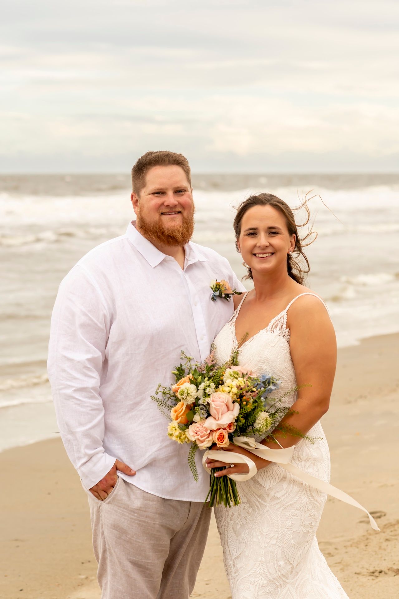 A couple poses together at the beach on their wedding day with a beautiful floral bouquet.