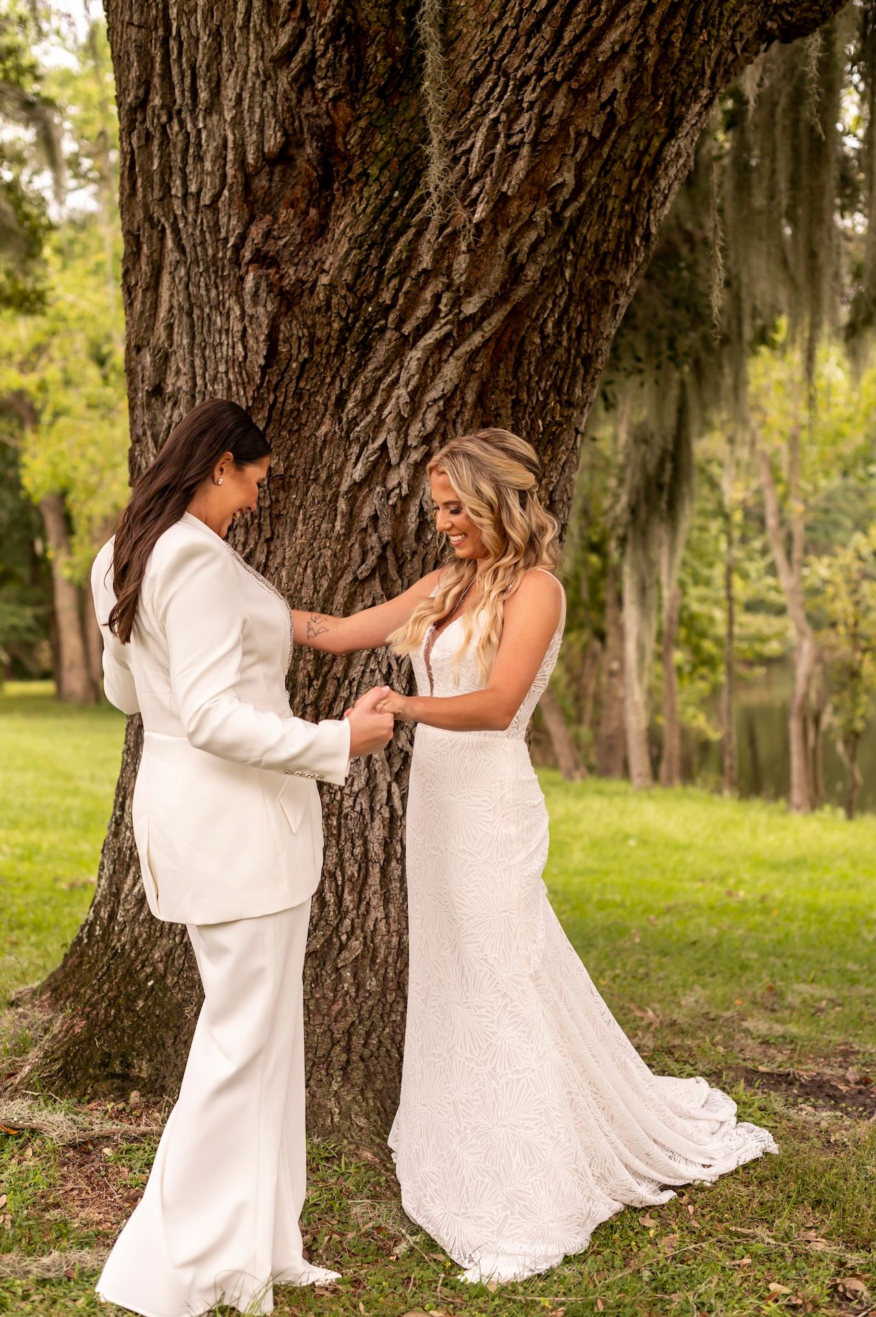 Two people in elegant white outfits share a joyful moment together under a large oak tree in a sunlit outdoor setting.