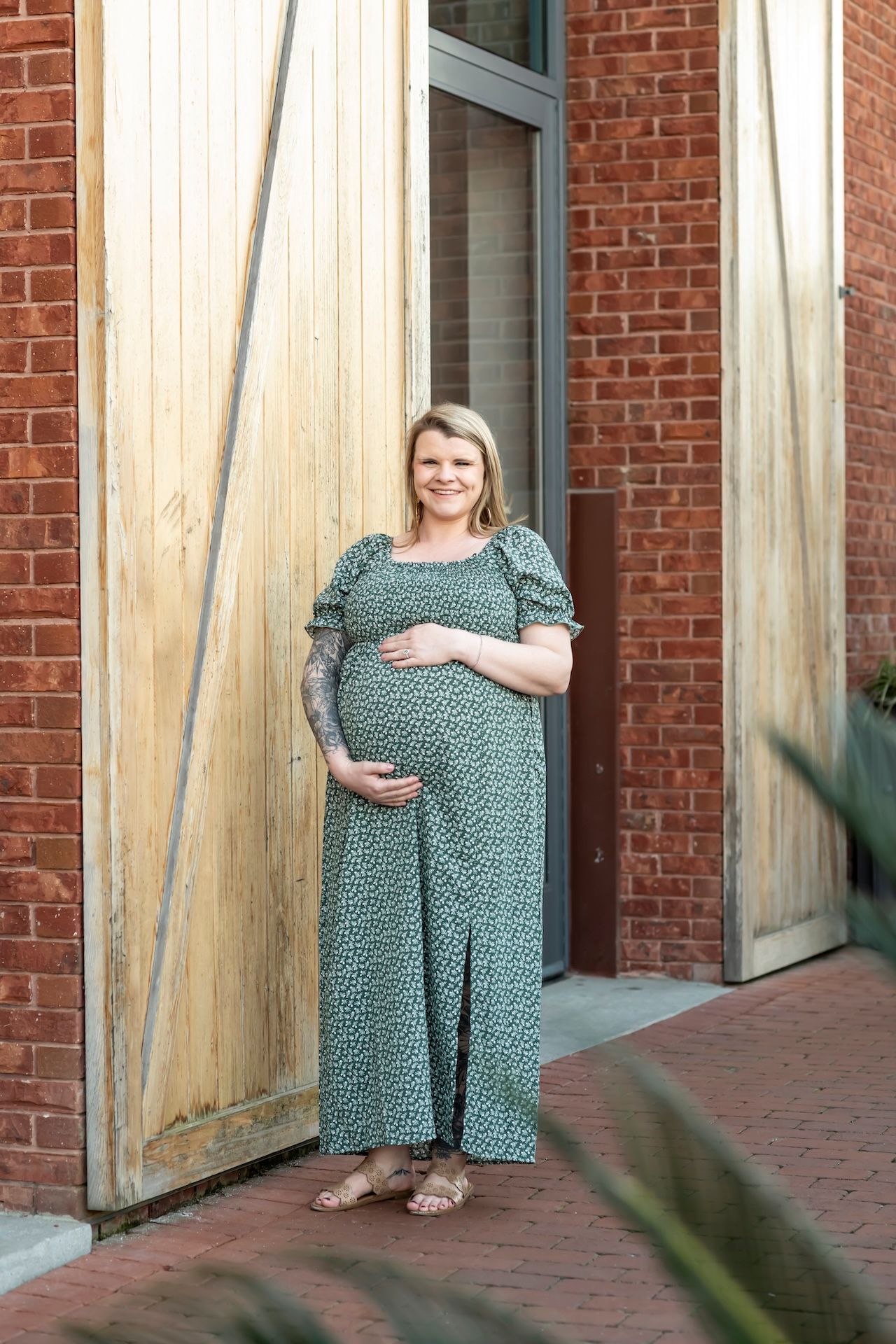 A person in a green patterned dress poses against a rustic wooden door beside a red brick wall.