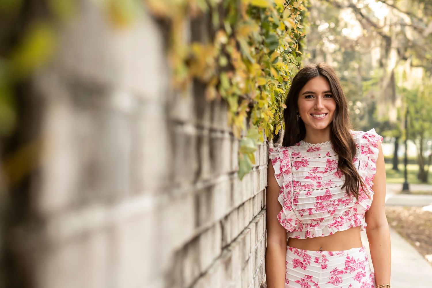 Someone wearing a pink floral two-piece outfit poses by a stone wall with greenery in an outdoor setting.