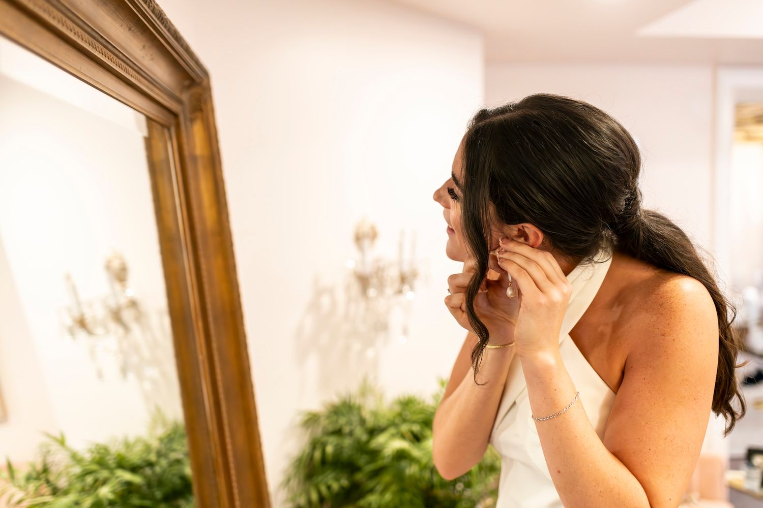 A person in a white wedding dress puts on earrings while looking in a mirror decorated with vines.