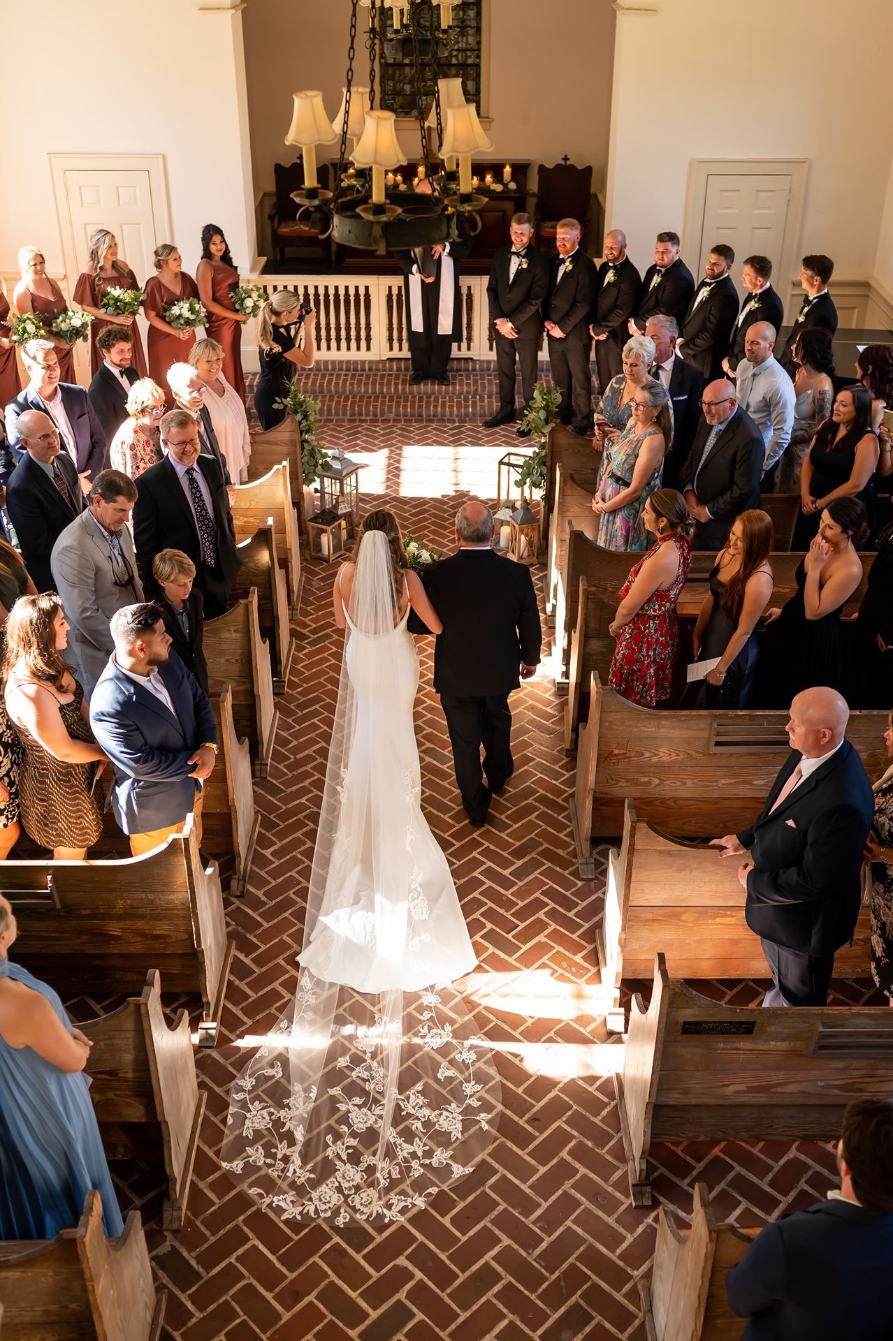 Wedding ceremony in a church with sunlight streaming through windows onto the brick aisle pattern as guests look on.