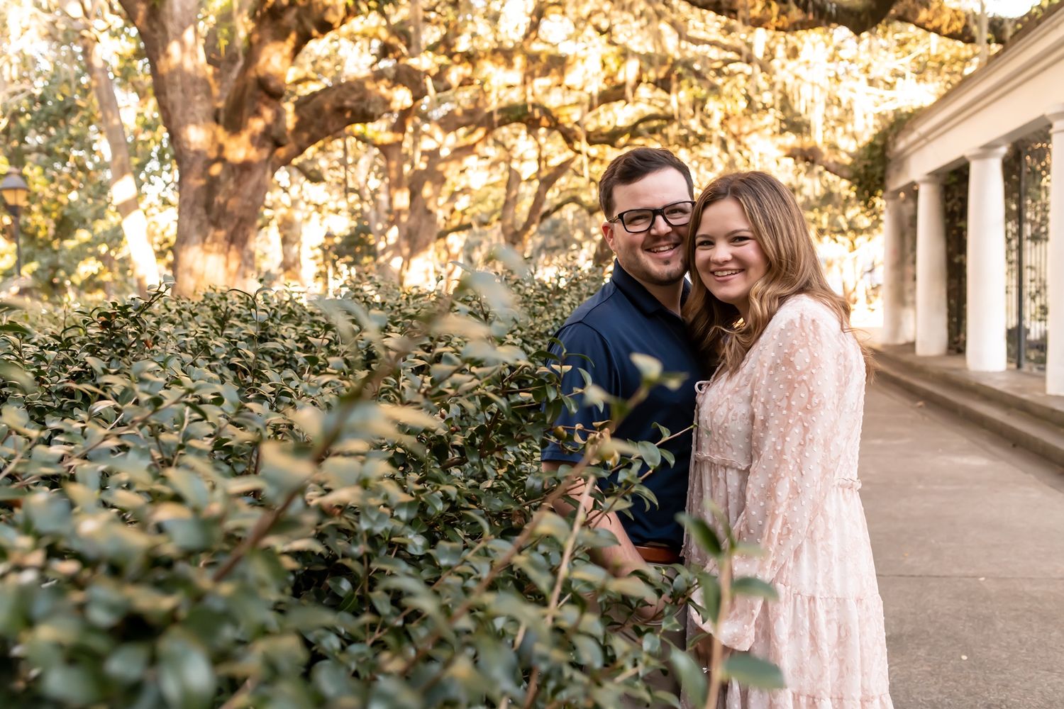 A romantic couple poses together near lush greenery and historic architecture during golden hour.