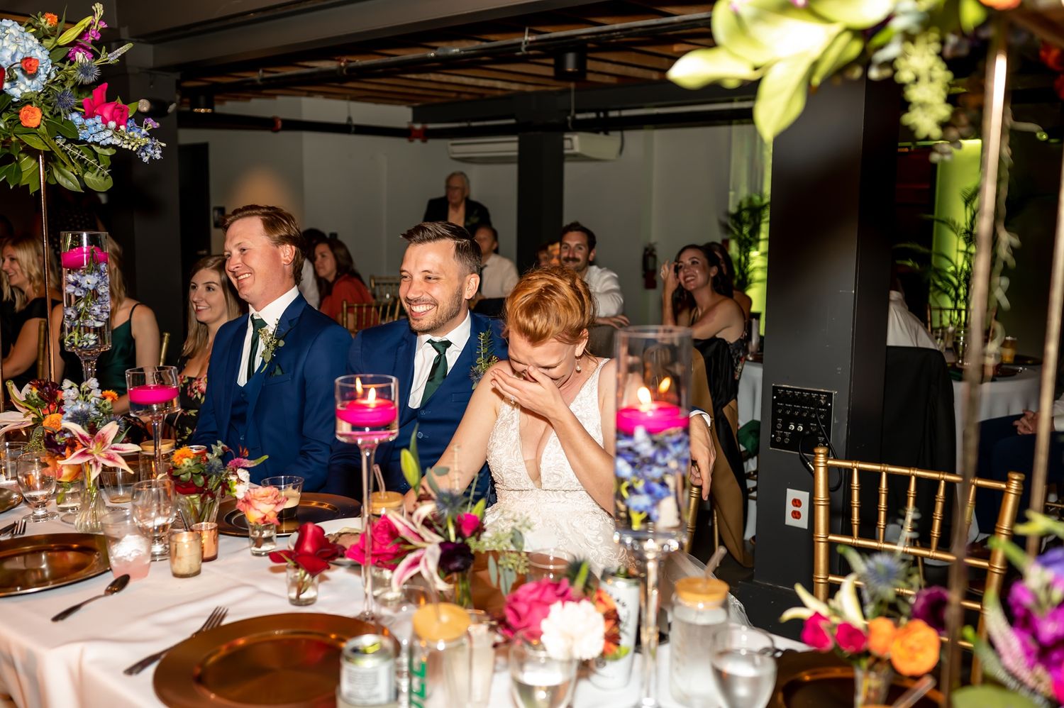 Wedding reception guests seated at an elegant table with vibrant pink floral centerpieces and gold decor.
