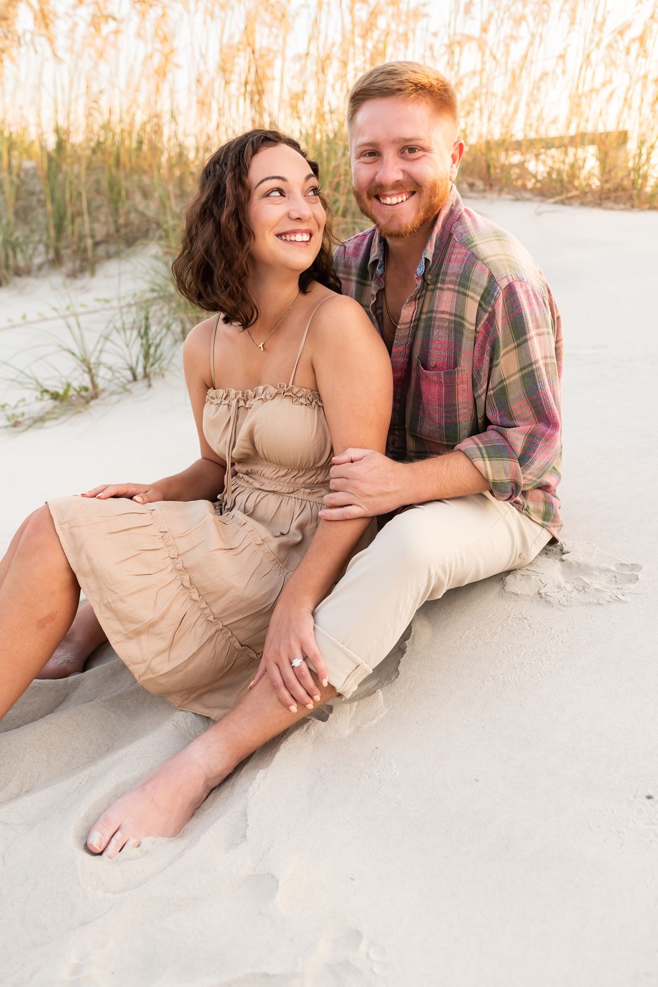 A couple sits together on a sandy beach at sunset with sea oats in the background.