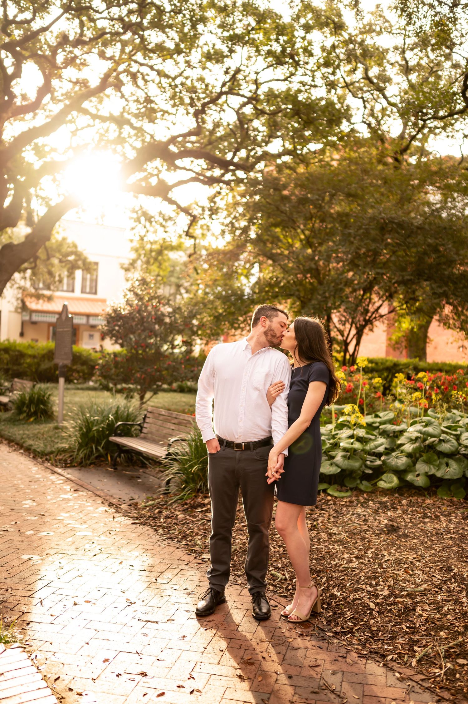 A couple enjoys a romantic moment on a garden path during golden hour with sunlight filtering through trees.