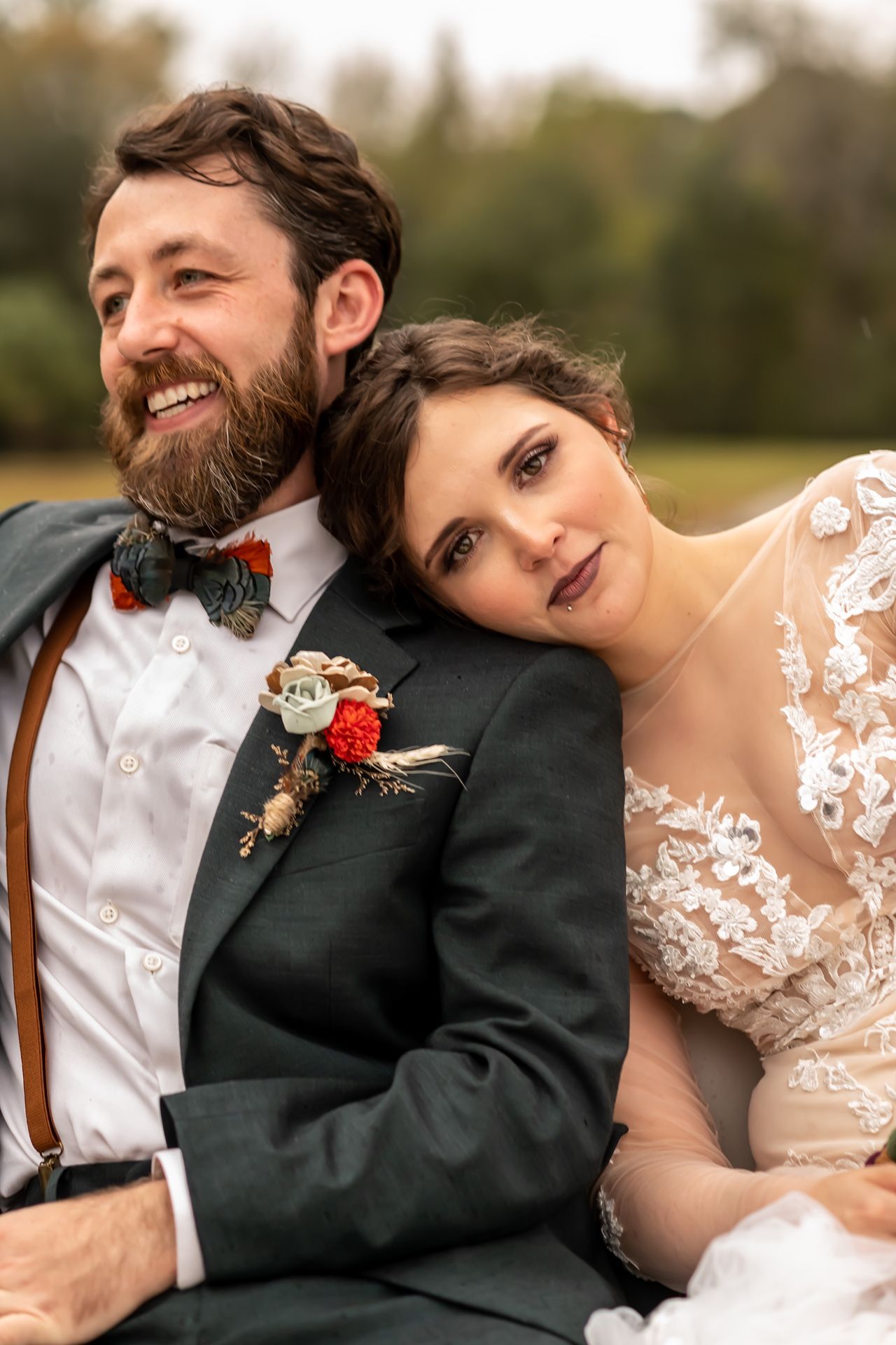 Newlyweds in wedding attire share tender moment outdoors, with groom in bowtie and suspenders and bride in lace dress.