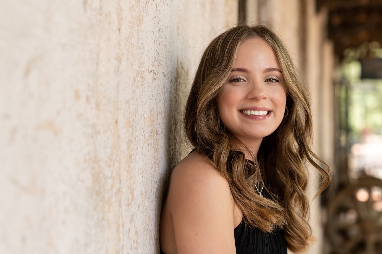 A person with long wavy brown hair and a bright smile poses in a natural outdoor setting with white columns.