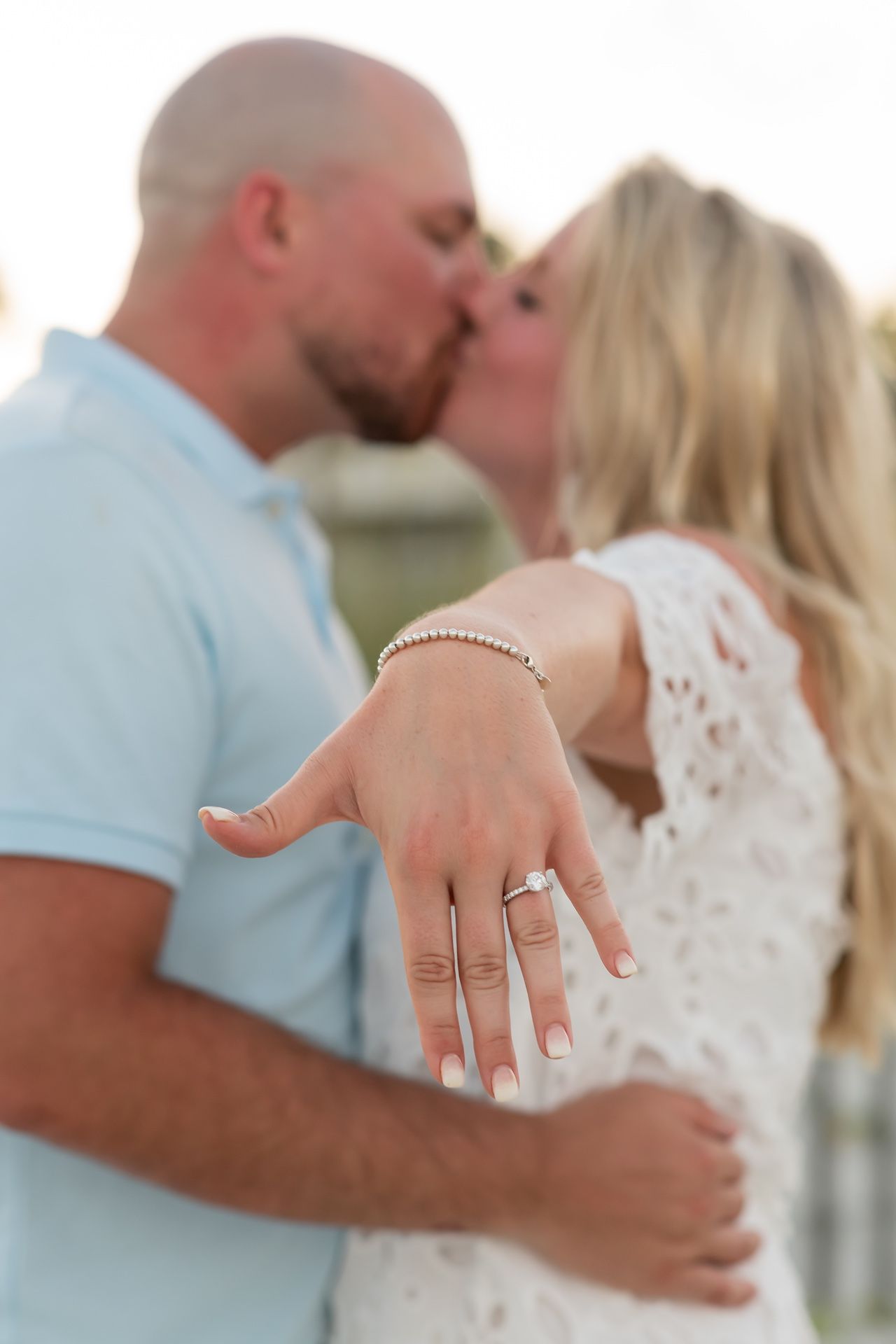 A woman showing off her engagement ring while sharing a romantic kiss in casual outdoor attire.
