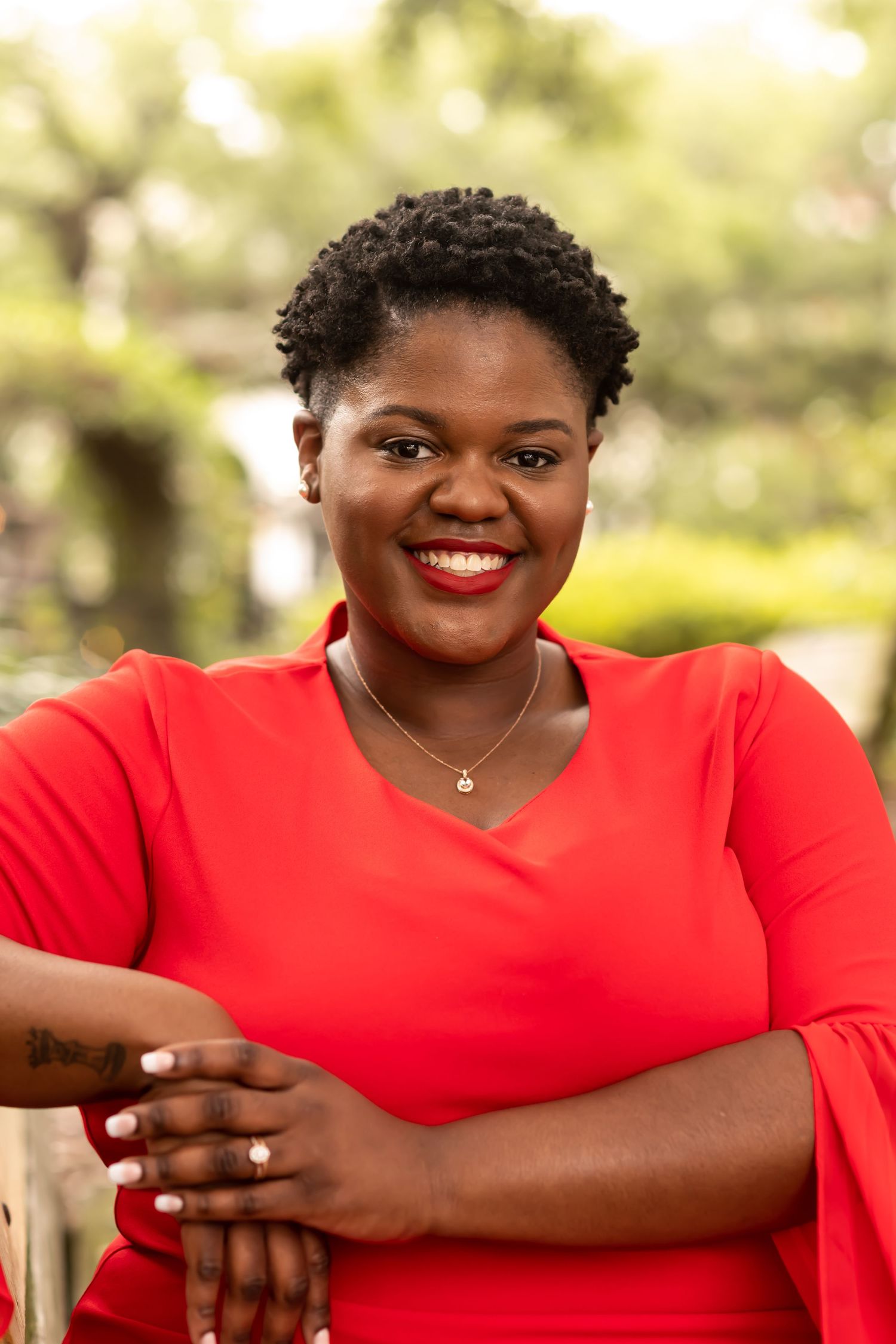 A warm and joyful portrait outdoors with a red V-neck top and delicate necklace against a soft, blurred background.