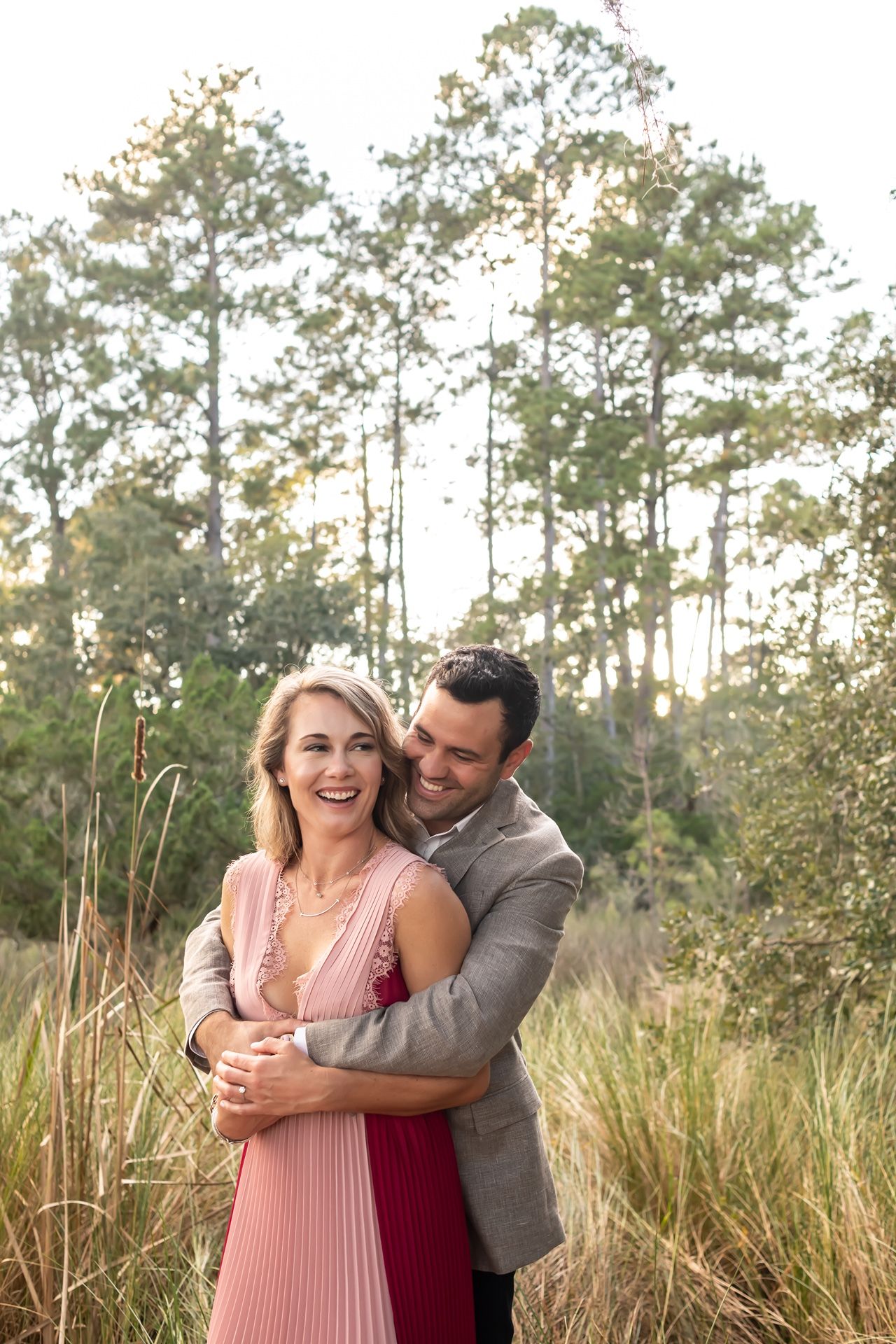 Joyful couple embraces and laughs together during outdoor photoshoot with tall pine trees in the background.
