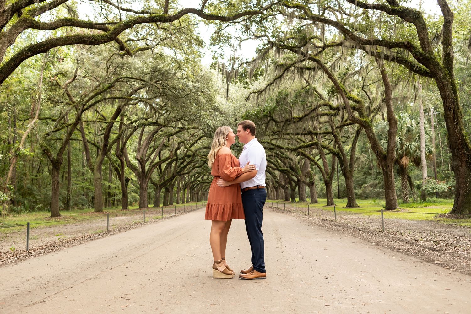 A couple walks down a scenic dirt path lined with massive oak trees draped in Spanish moss.