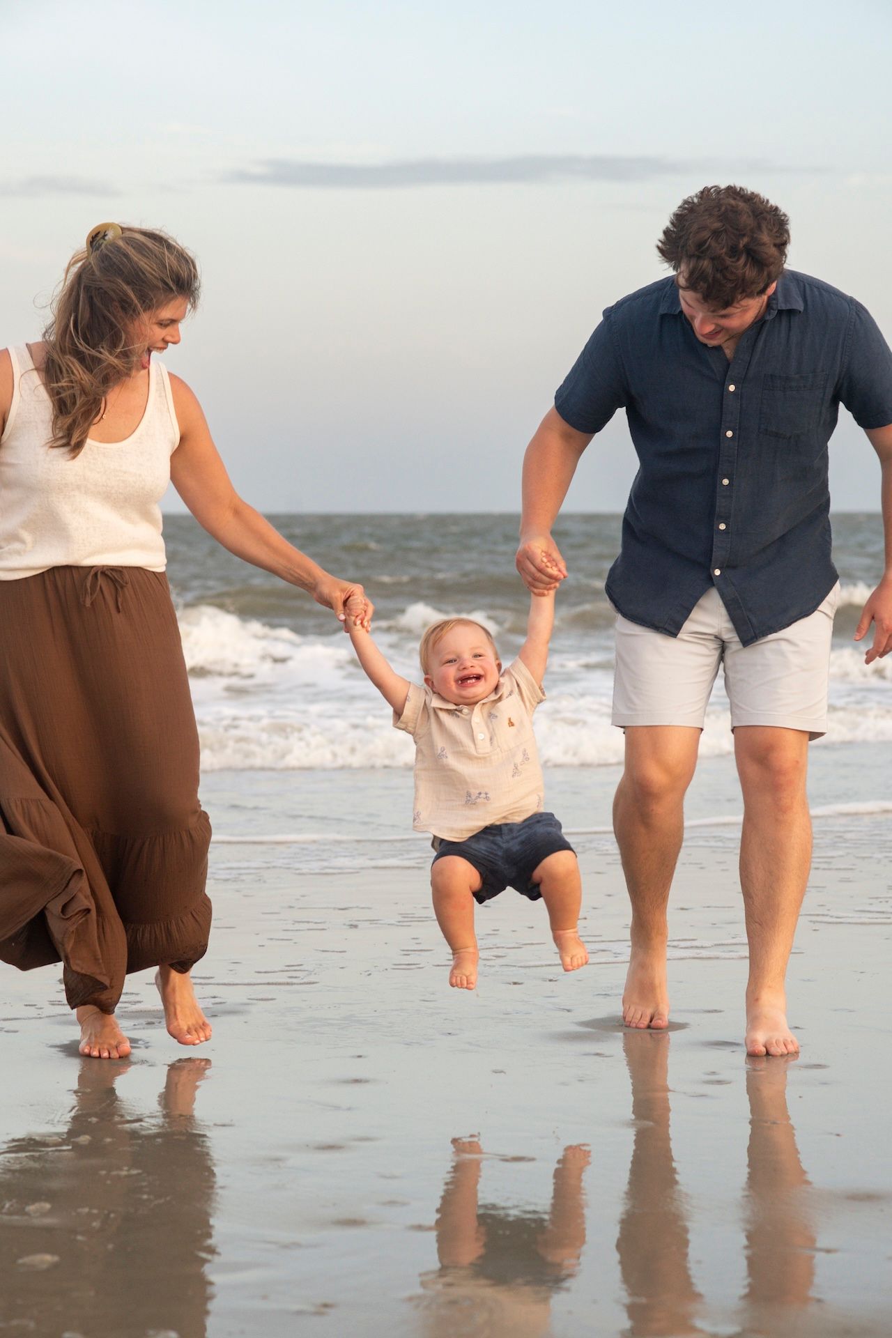 Happy baby laughing as parents swing child on sunny beach, family enjoying summer vacation by ocean waves.