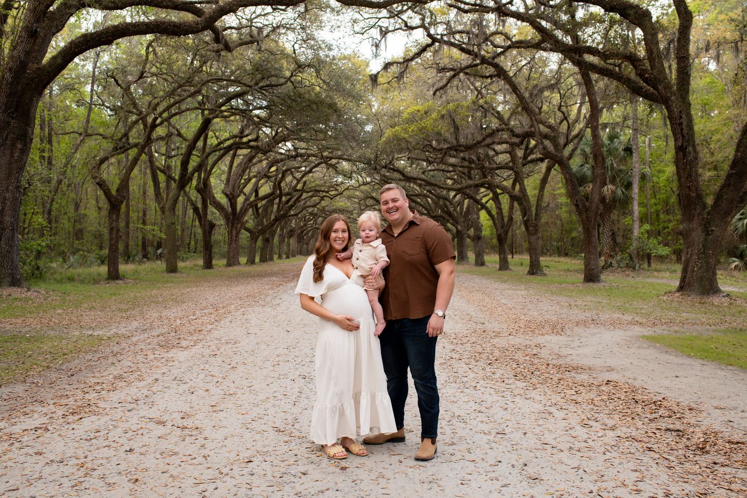 Pregnant woman in white dress with husband and toddler posing on tree-lined path for maternity family photo.