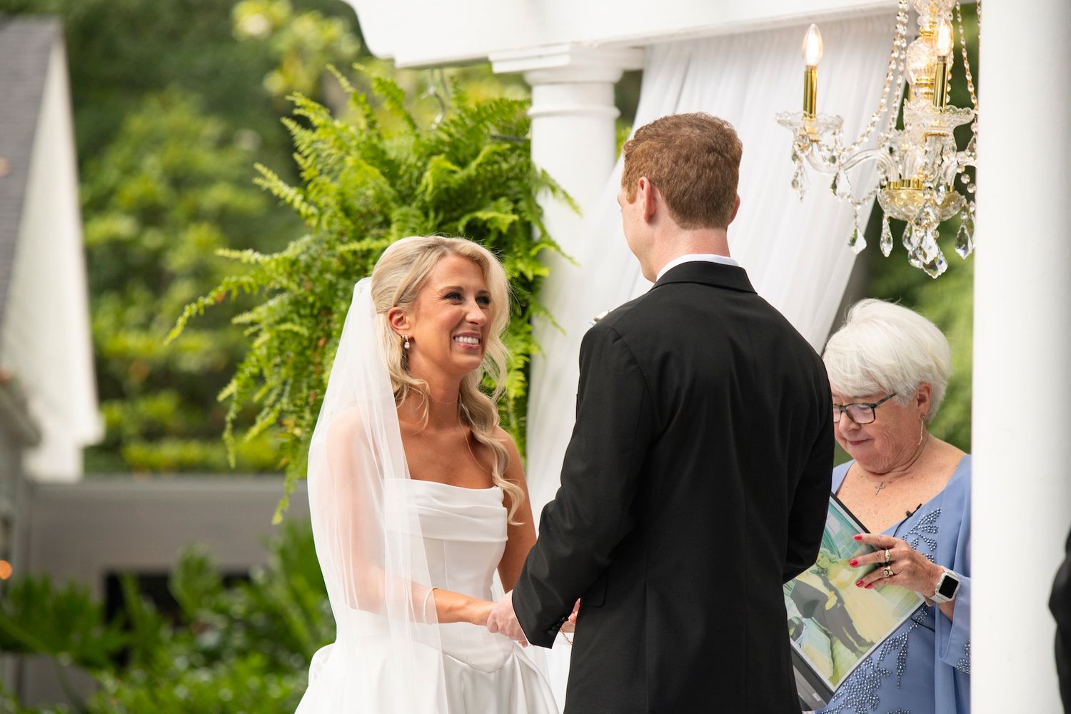 Bride smiling joyfully at groom during outdoor wedding ceremony with chandelier and lush greenery backdrop.
