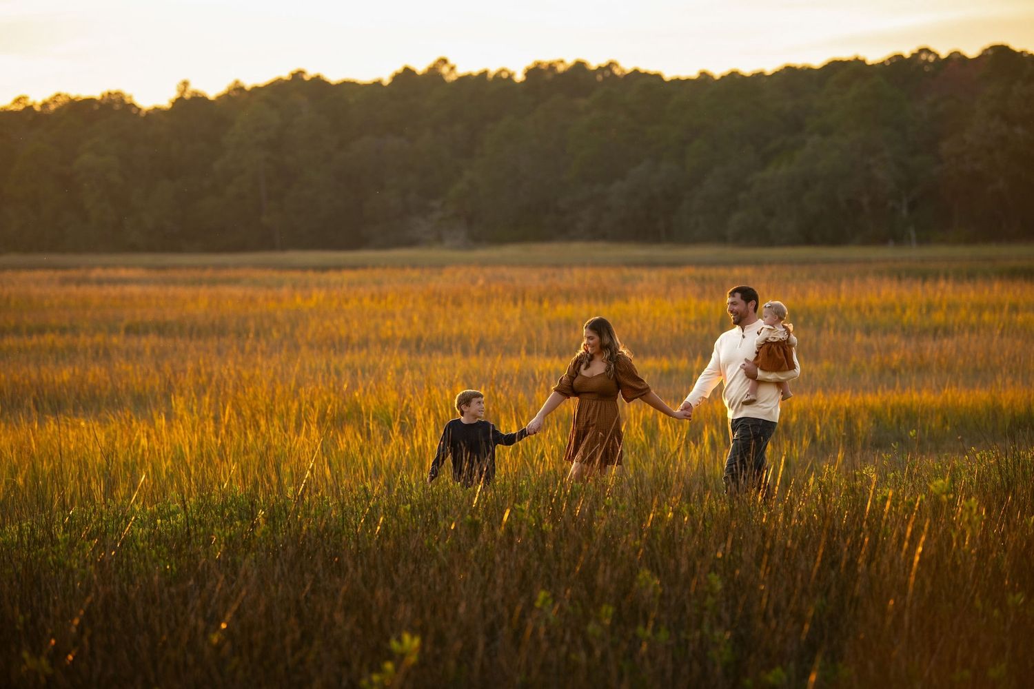 Family walking hand-in-hand through golden marsh field at sunset with trees in background.