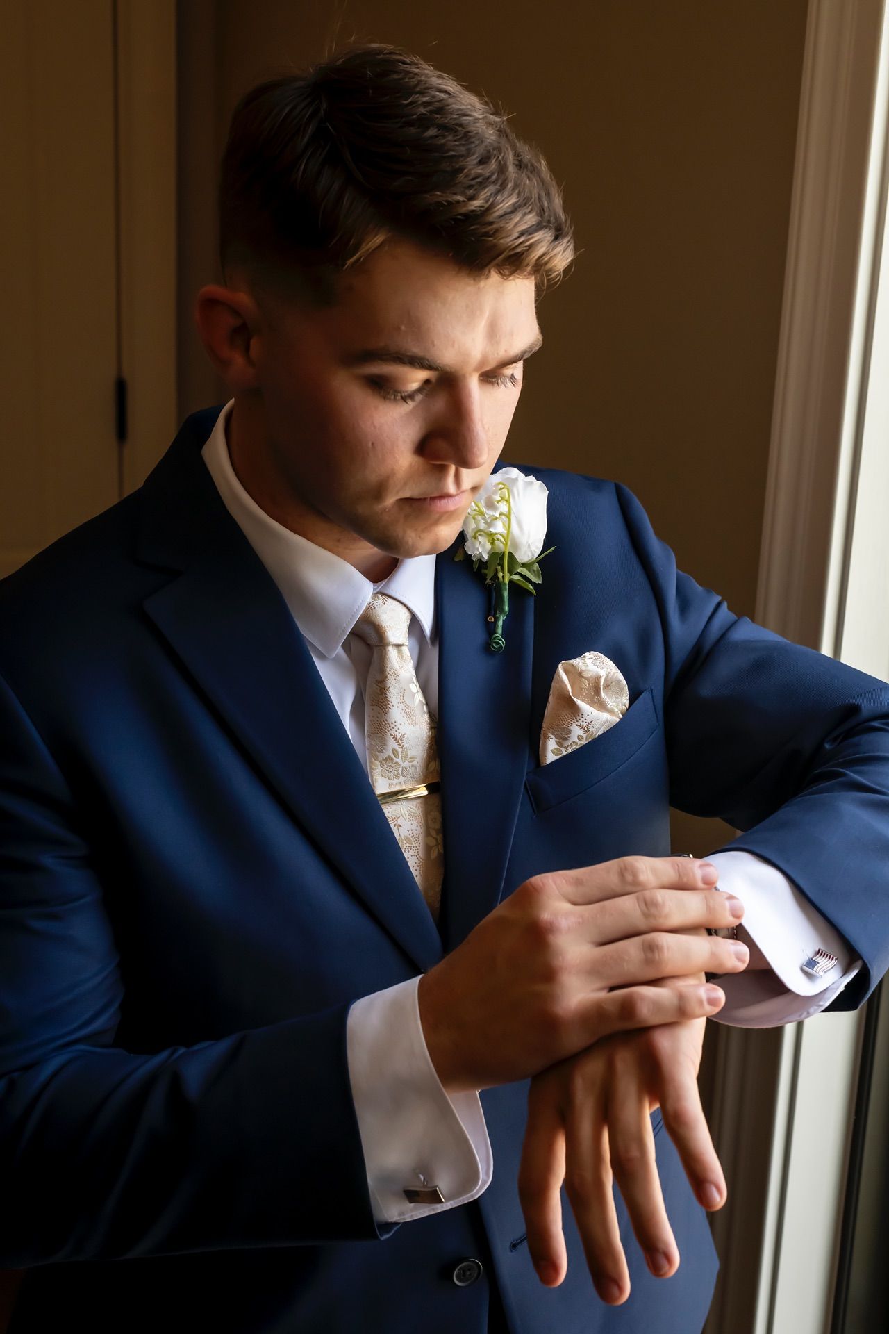 A groom in a navy suit adjusts his cufflinks while standing near a window.