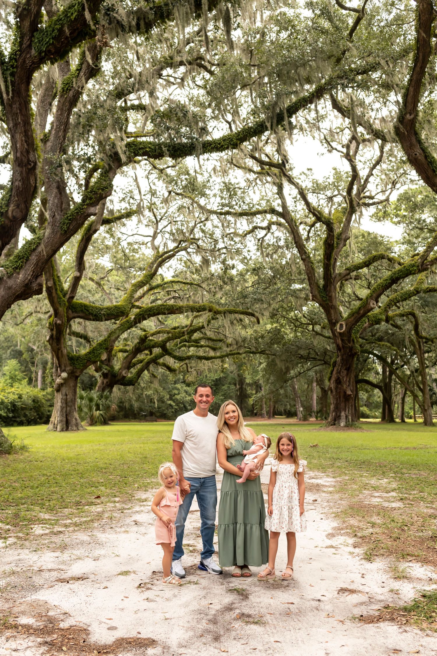 A family shares a candid moment together on a sandy path beneath towering oak trees draped in Spanish moss.