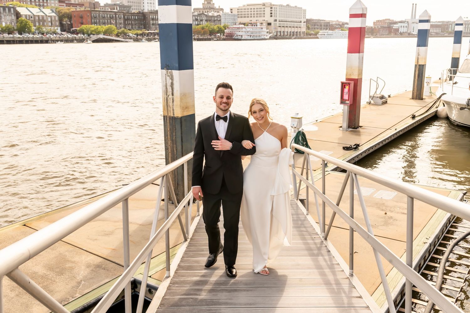 Bride and groom walking hand-in-hand on a waterfront dock, smiling, with a harbor and city skyline in the background.