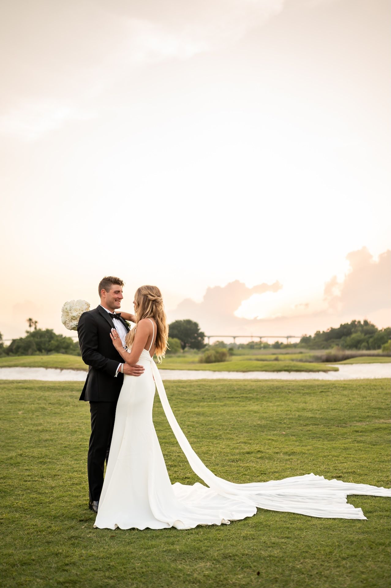 Bride and groom embrace at sunset on a golf course, bride wearing a sleek white gown with long train.