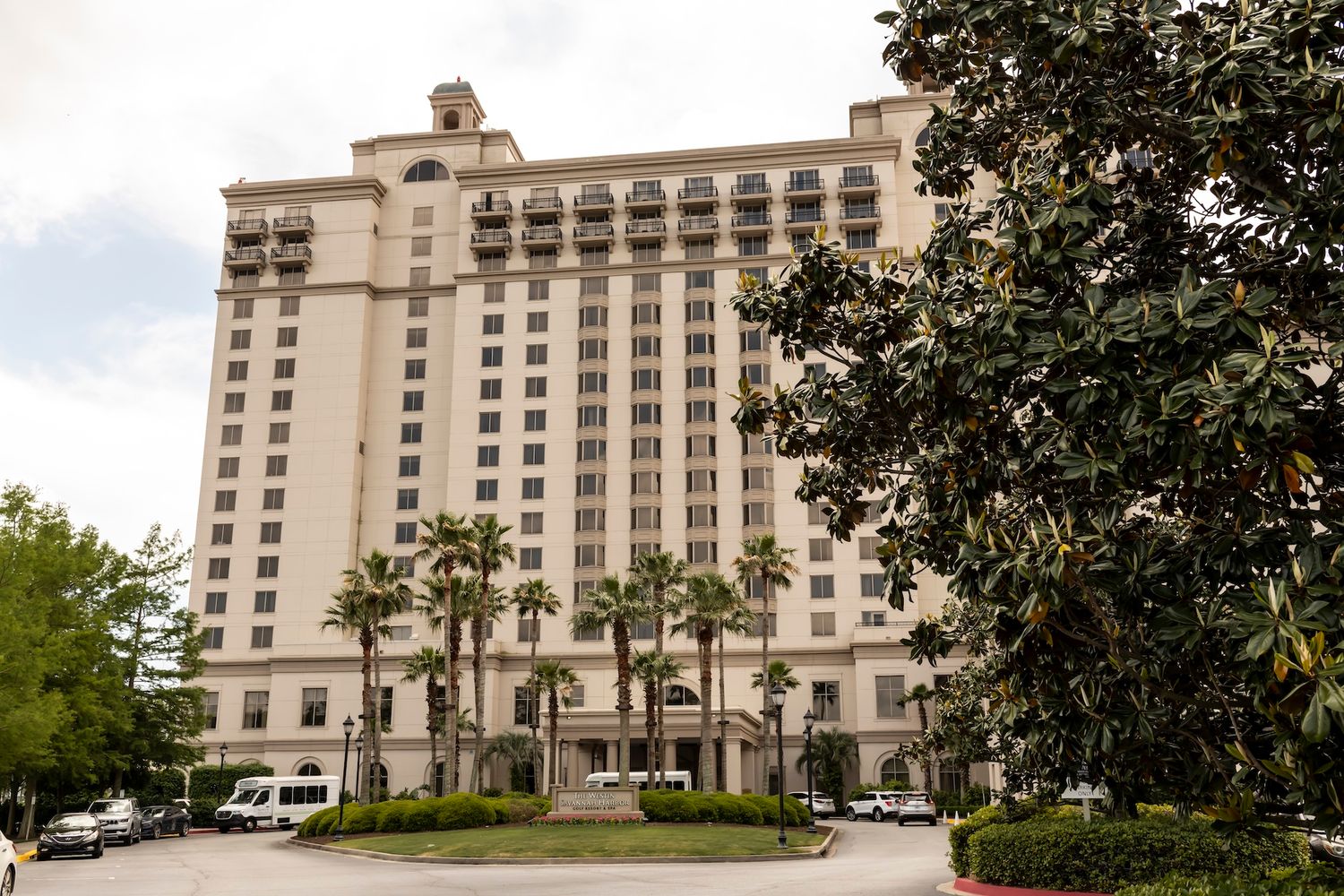 Large luxury hotel with cream facade, palm trees, and circular driveway surrounded by lush greenery under cloudy skies.