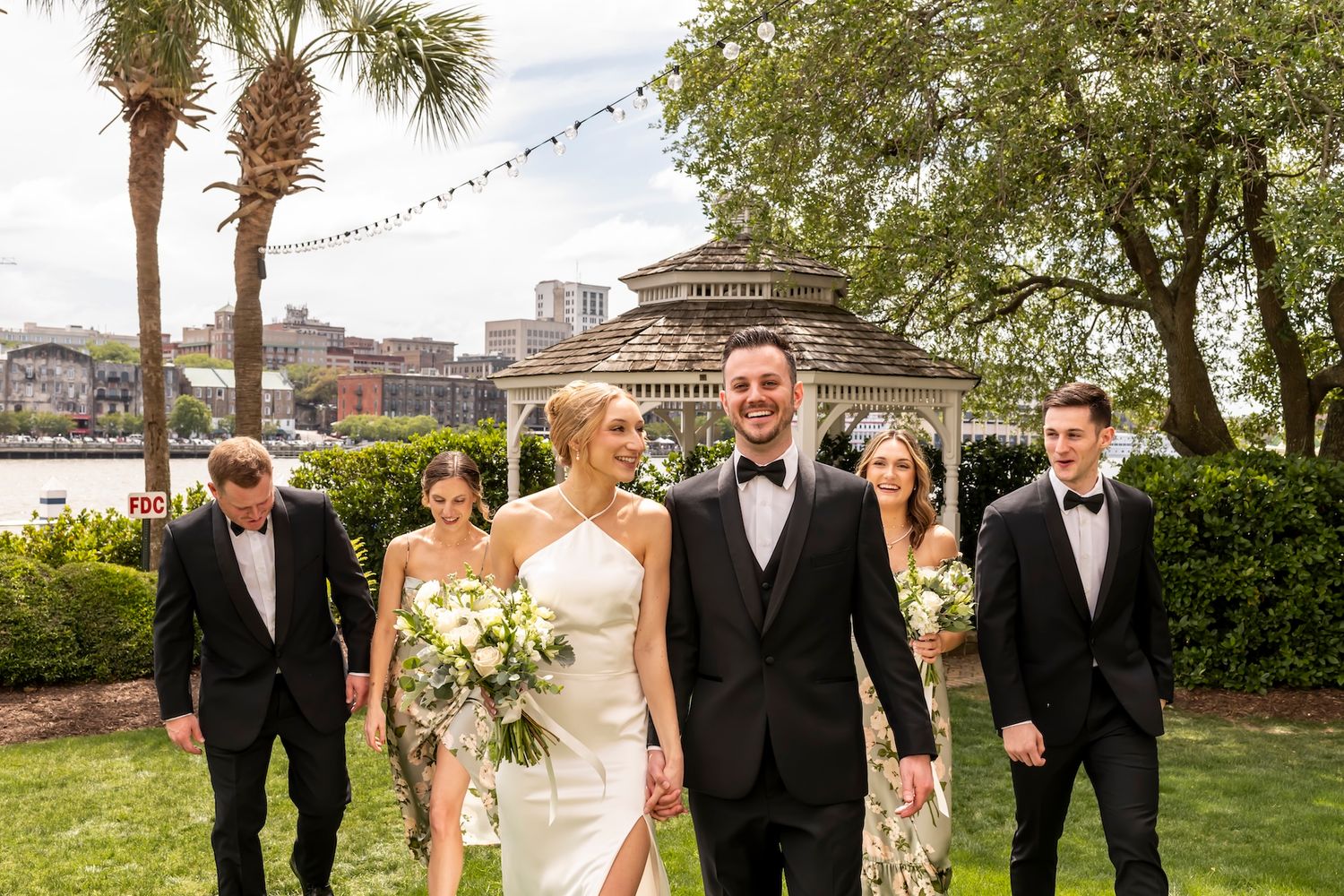 Wedding party walks outdoors near waterfront gazebo with bride in halter gown and groom in black tuxedo.