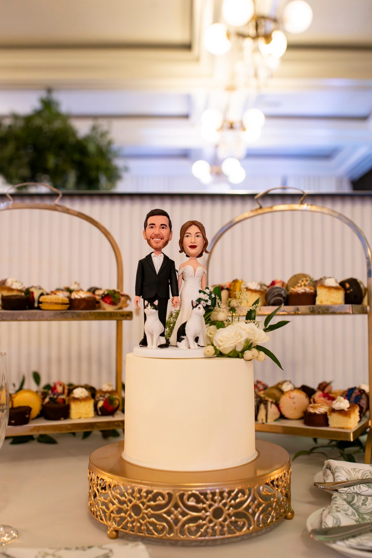 White wedding cake with custom topper displayed on gold decorative stand with dessert table in background.