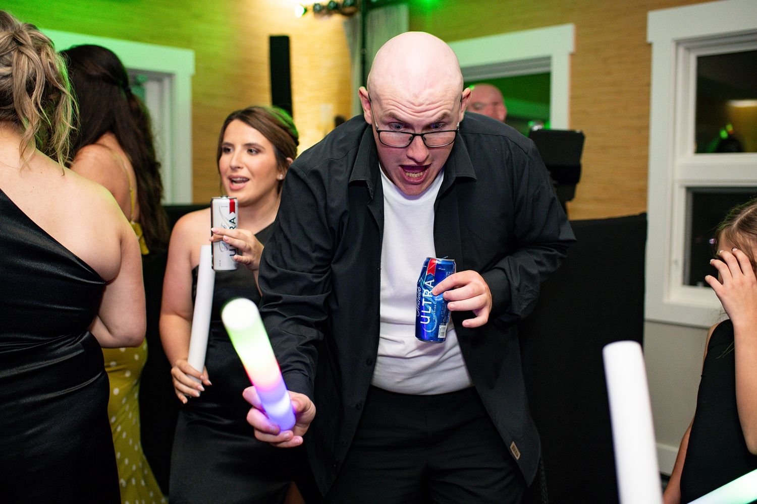 Party guest in a black suit dances energetically with glow sticks at a celebration event.