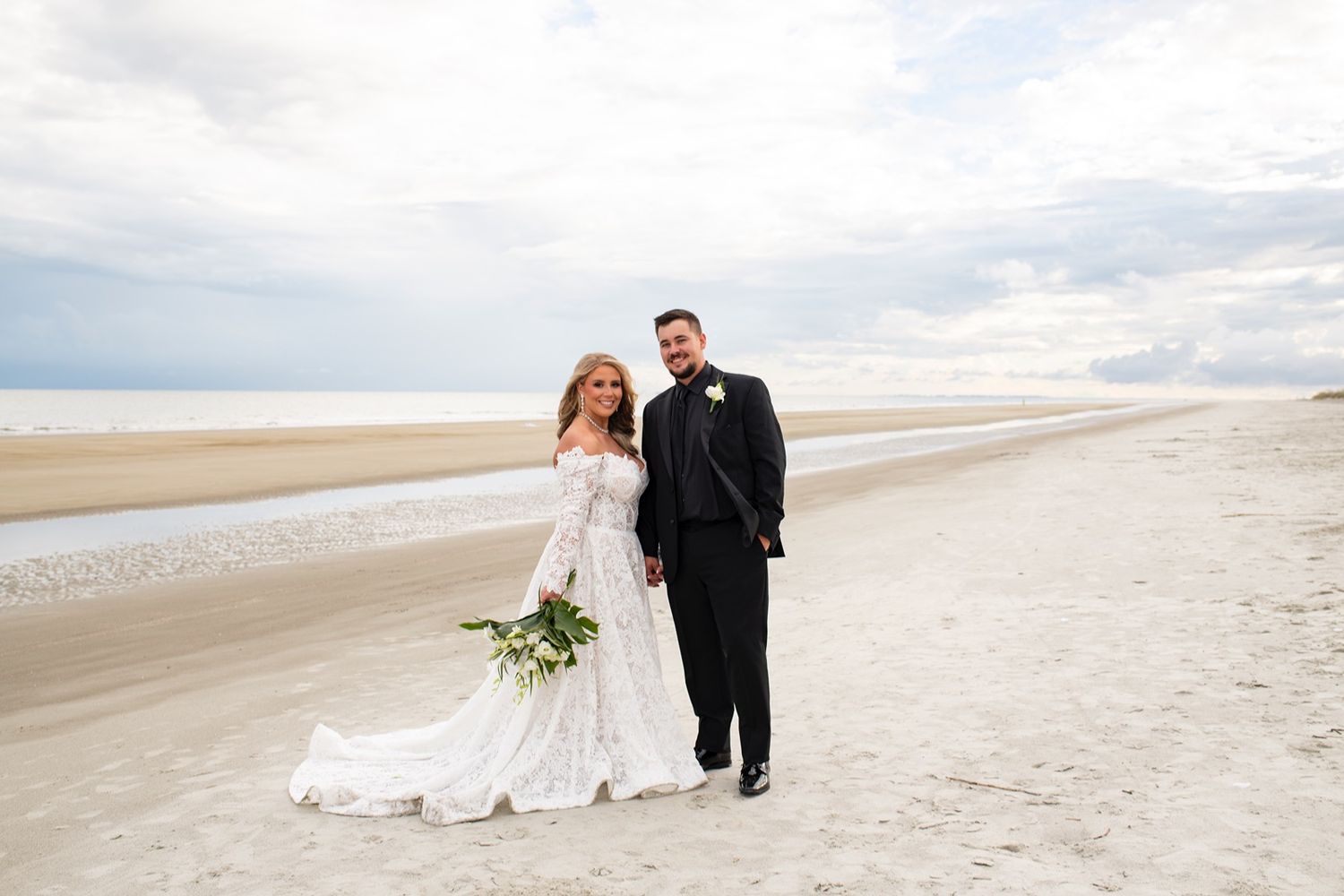 A couple in wedding attire walks along a scenic beach shoreline under cloudy skies.