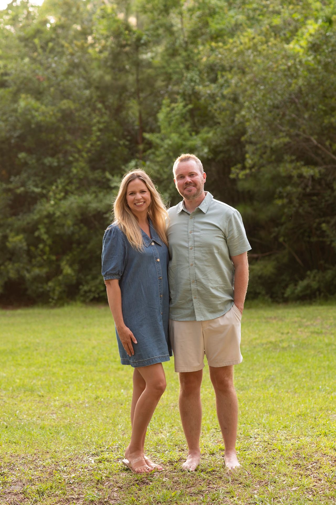 A smiling couple stands barefoot on green grass outdoors, woman in denim dress, man in sage shirt and khaki shorts.