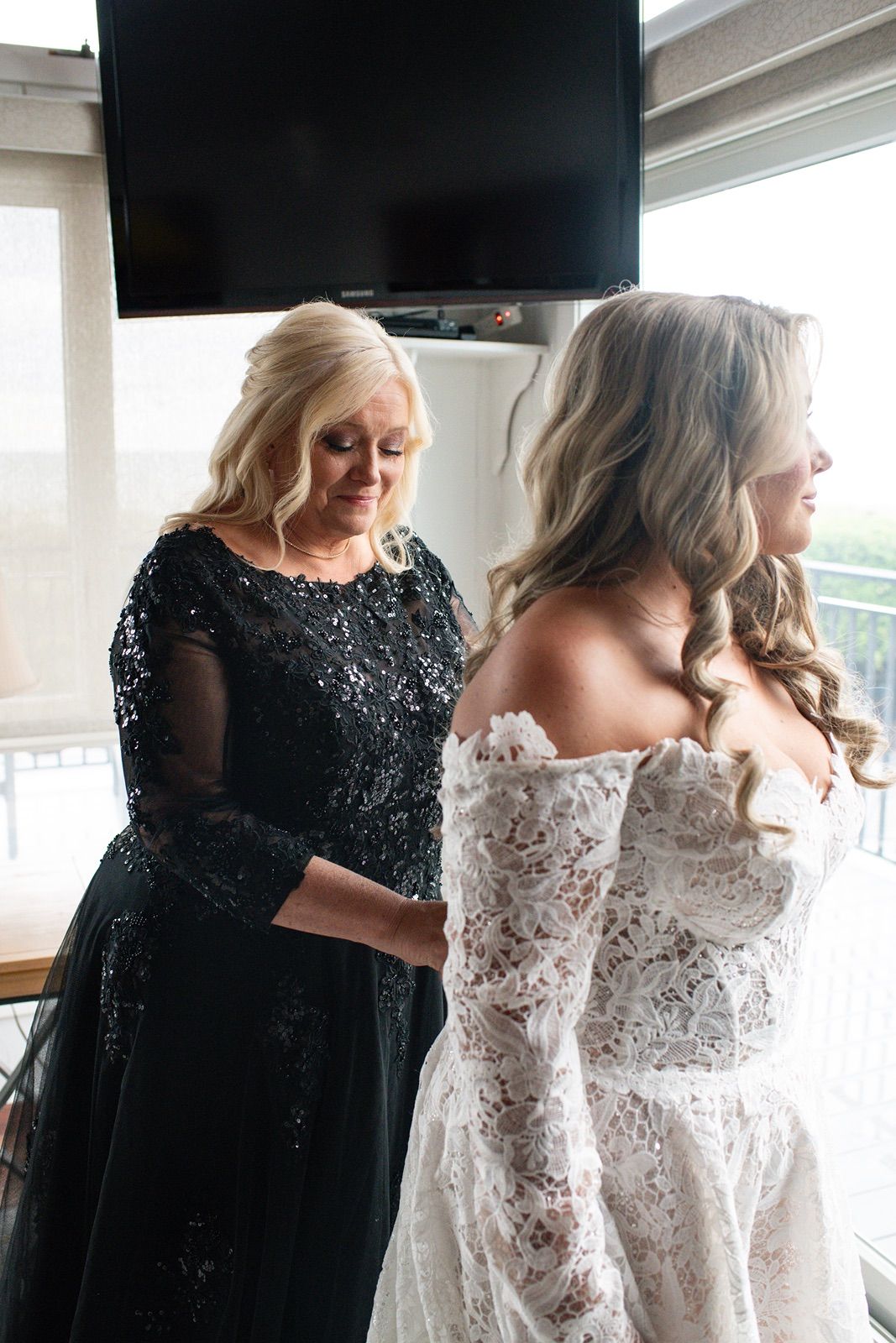 A woman in a black sequined dress helps adjust a white lace wedding gown during bridal preparations.